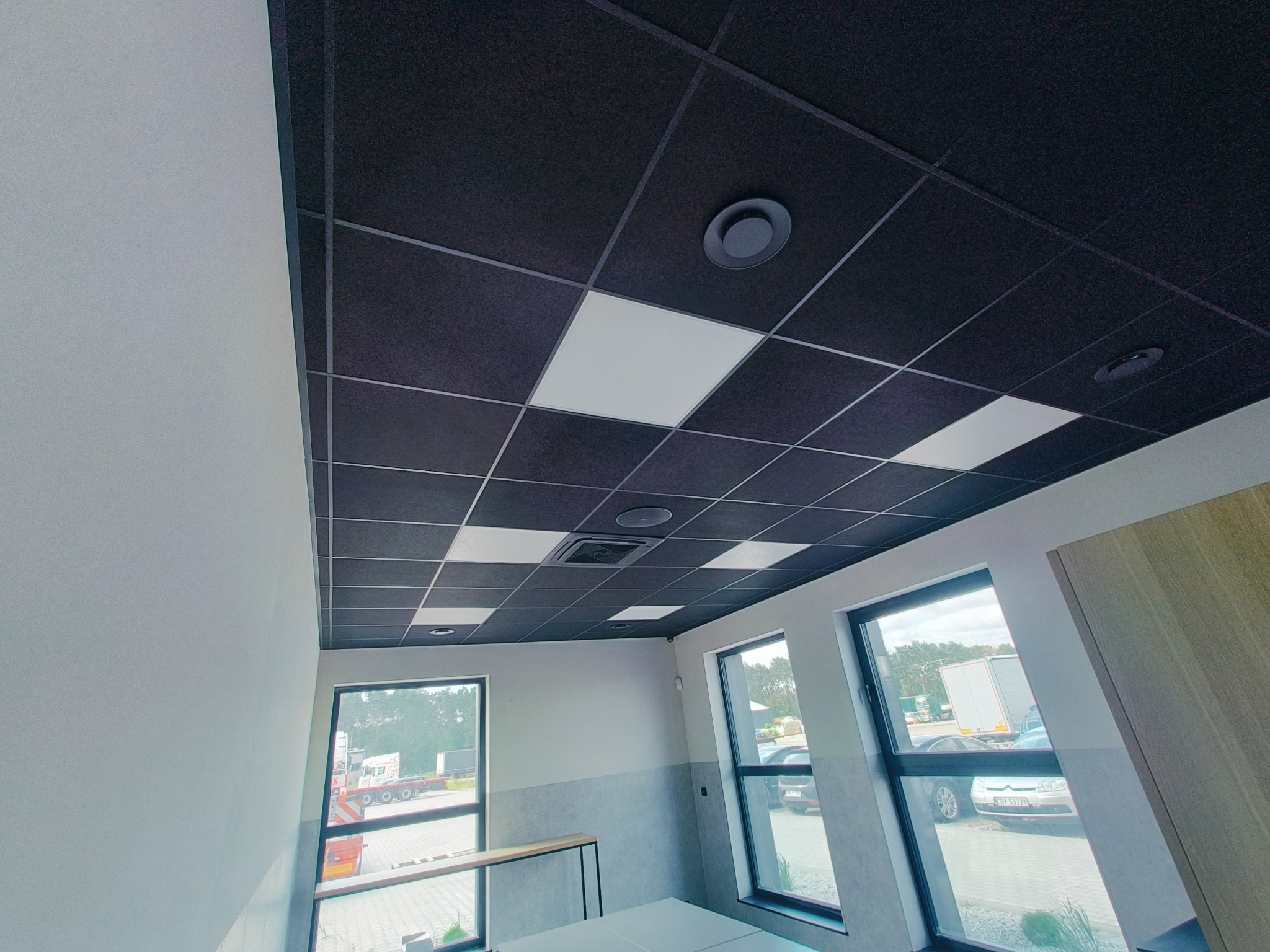 Czarny sufit kasetonowy z wbudowanym oświetleniem LED i okrągłymi lampami. Widok na jasne ściany i okna z widokiem na zewnątrz. Nowoczesny design.