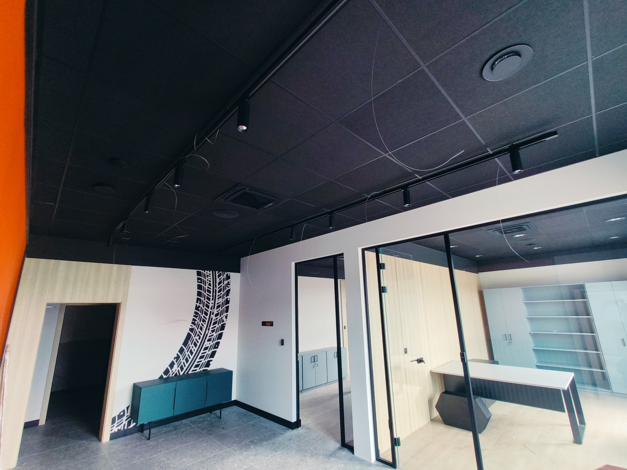 Nowoczesne biuro po remoncie: czarny sufit z oświetleniem, ściana z motywem opony, turkusowa szafka i przeszklone biuro. Widok na wnętrze przez otwarte drzwi.