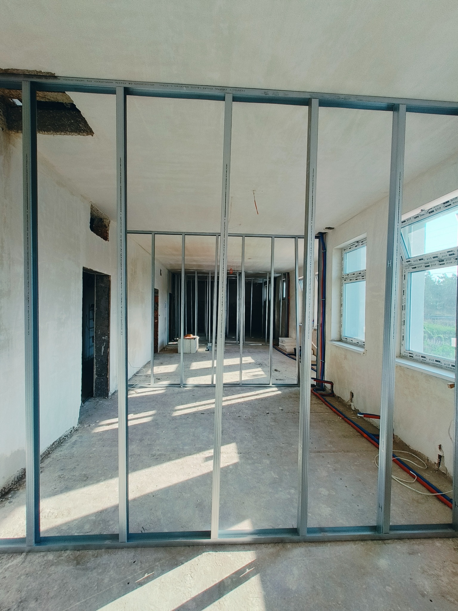 Metalowa konstrukcja szkieletowa ścian działowych w surowym wnętrzu. Widoczne instalacje i okna. Prace wykończeniowe w trakcie realizacji.