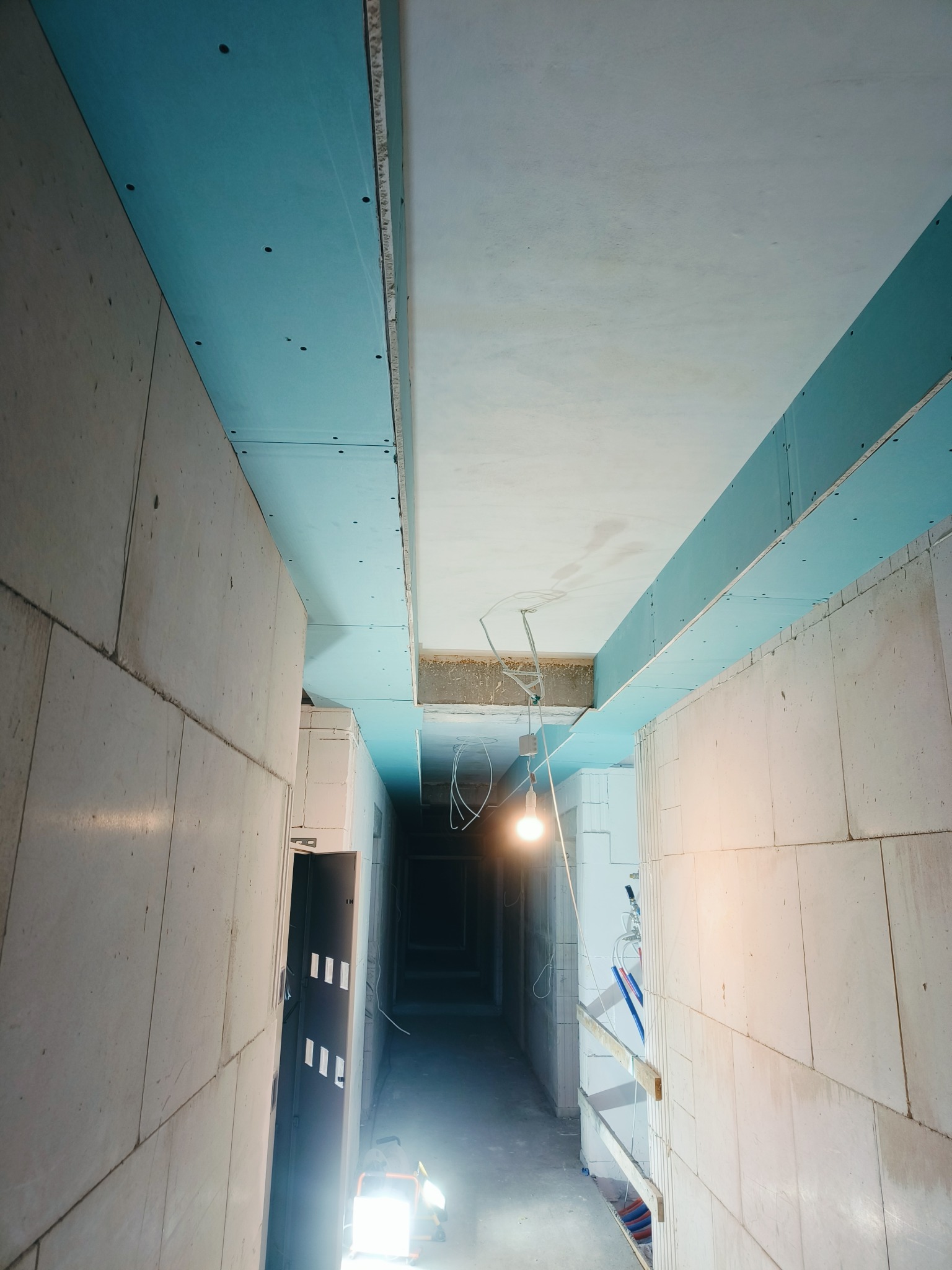 Korytarz w trakcie prac wykończeniowych: ściany z bloczków, zamontowane płyty karton-gips na suficie, oświetlenie robocze. Widok w perspektywie.