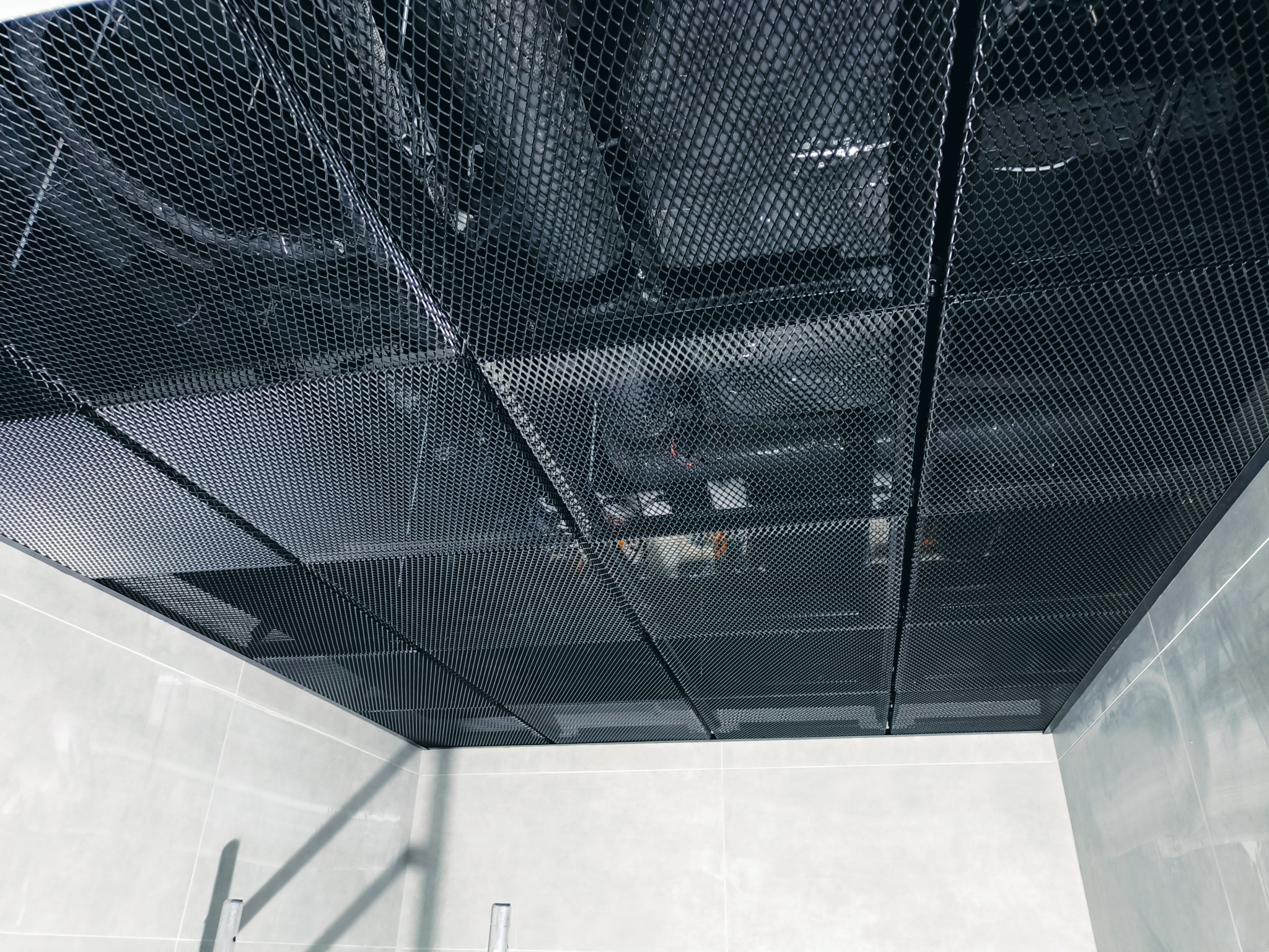 Czarny sufit podwieszany z metalowej siatki w pomieszczeniu z płytkami. Widoczne instalacje nad sufitem.