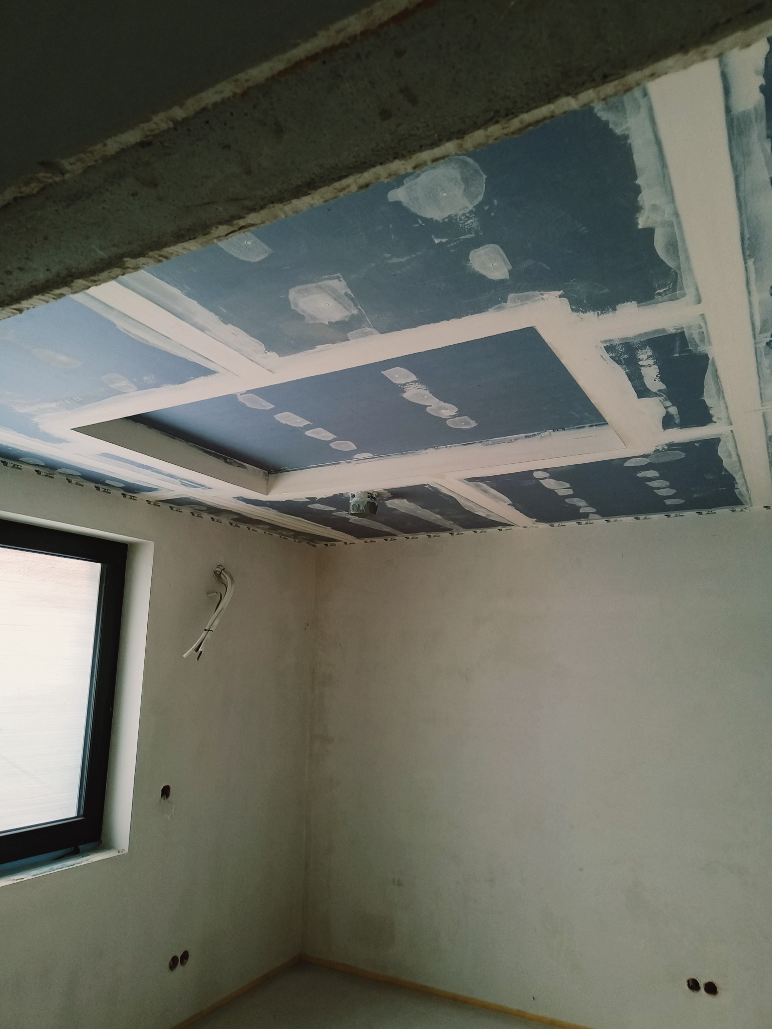 Montaż płyt gipsowo-kartonowych na suficie z widocznymi łączeniami. Widoczne przygotowania do szpachlowania. W tle okno i kable elektryczne.