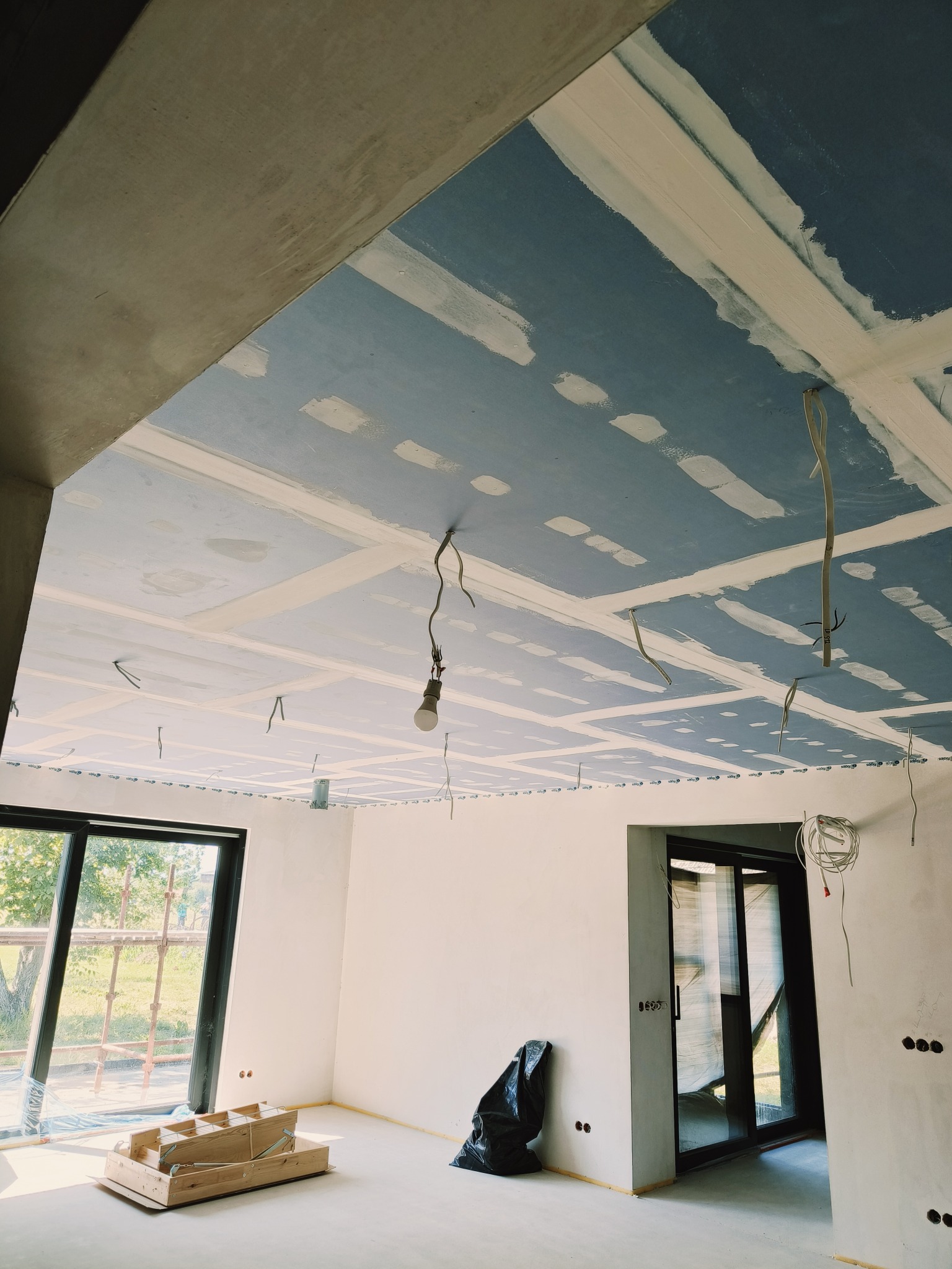 Wnętrze w trakcie prac: sufit z płyt karton-gips, widoczne łączenia i kable elektryczne. Na podłodze narzędzia i worek na śmieci, okno z widokiem na zewnątrz.