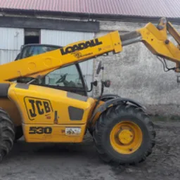 Żółta ładowarka teleskopowa JCB 530 podnosi na widłach duży, biały worek typu Big Bag na tle budynku gospodarczego.
