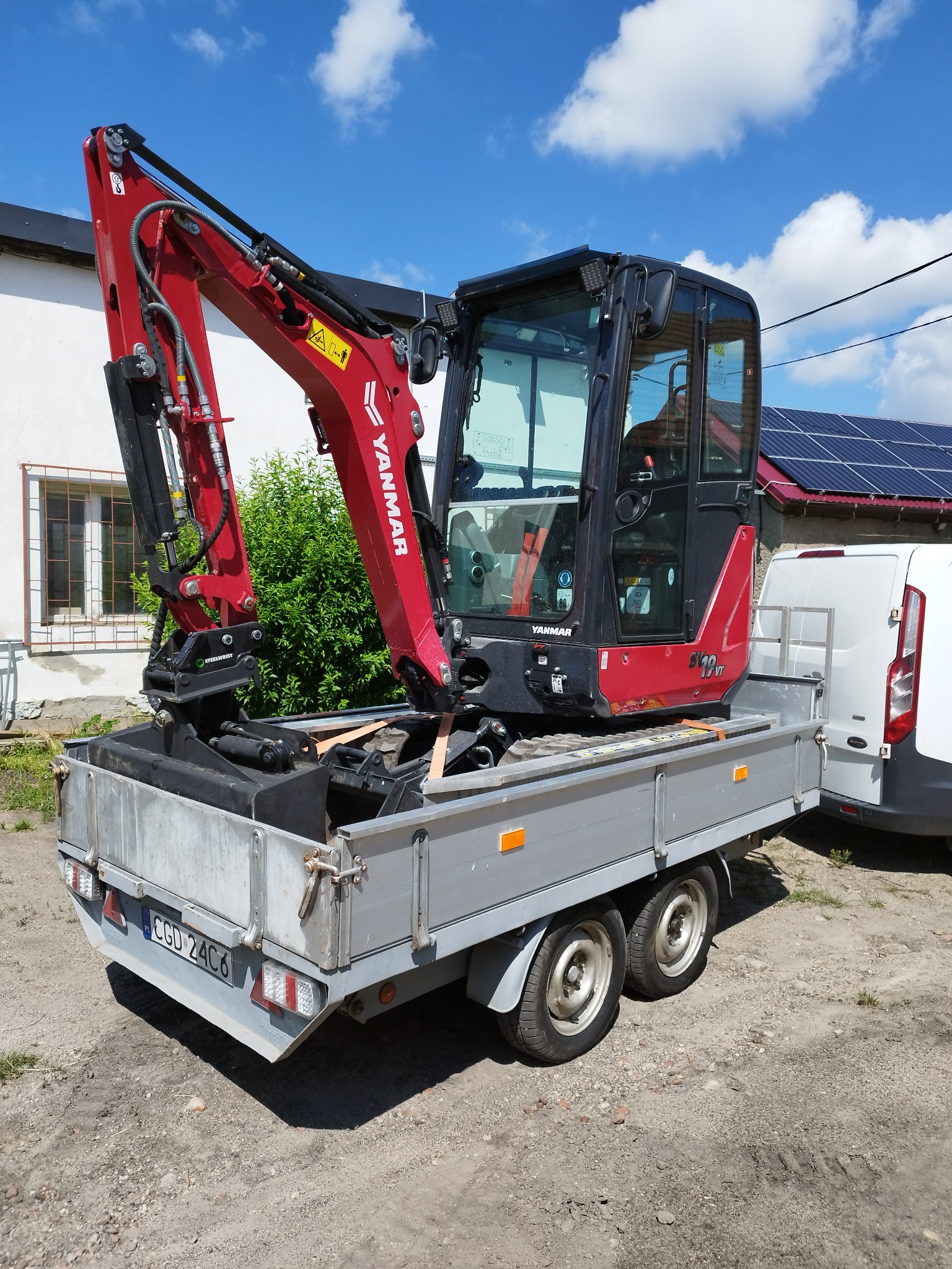 Czerwona mini koparka Yanmar 19 VT na szarej przyczepie dwuosiowej, zaparkowana obok białego busa, na tle budynku z panelami słonecznymi i fragmentem zieleni.