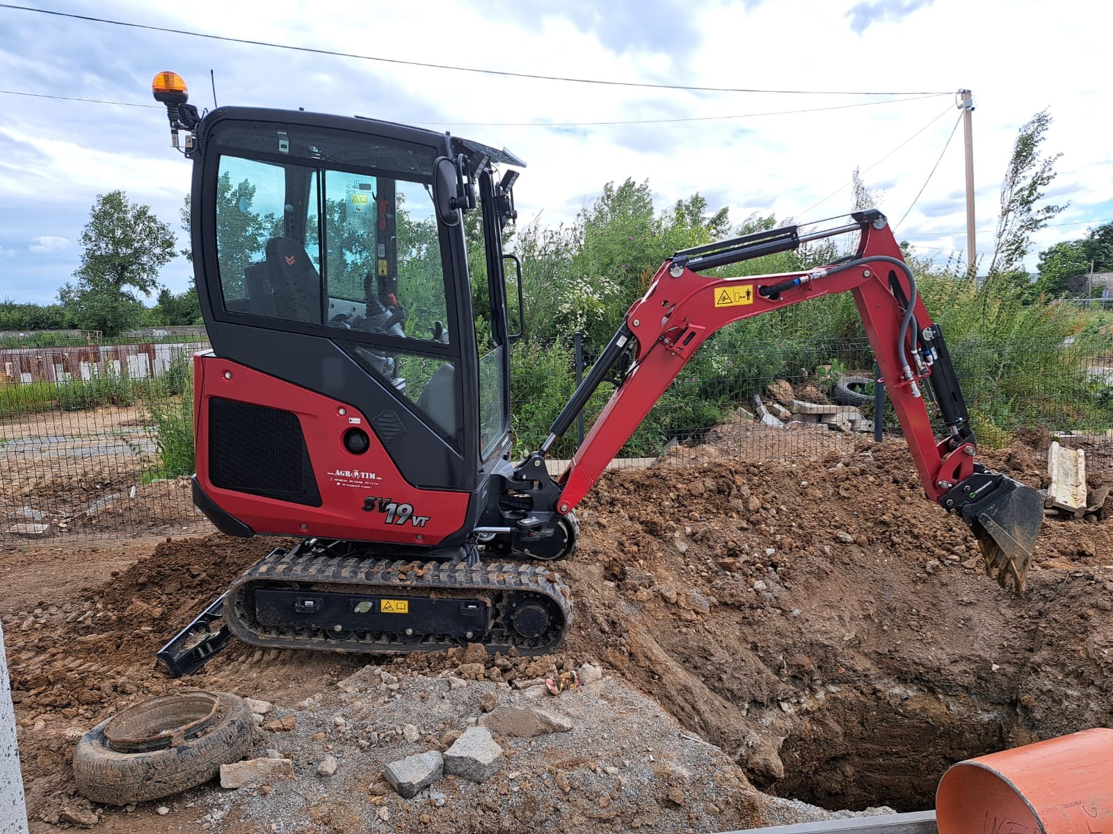 Czerwona mini koparka gąsienicowa SV19VT marki AGR-OTIM wykonuje prace ziemne, widoczny wykop z urobkiem, opona i rura PCV obok wykopu.