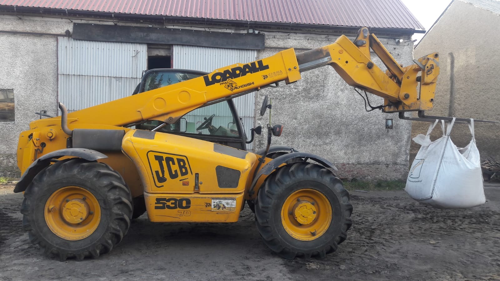 Żółta ładowarka teleskopowa JCB 530 podnosi na widłach duży, biały worek typu Big Bag na tle budynku gospodarczego.