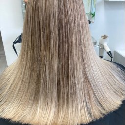 KANKOWSKA Salon Fryzjerski - Długie, proste włosy z koloryzacją sombre, przechodzące od ciemniejszego blondu u nasady do jaśniejszego na końcach, widoczne z tyłu, w tle suszarka i lustro.