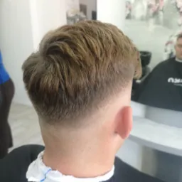 Męskie włosy po strzyżeniu z cieniowanym tyłem głowy, widoczny w lustrze fryzjer w czarnej pelerynie, białe ściany i dekoracje kwiatowe w tle.