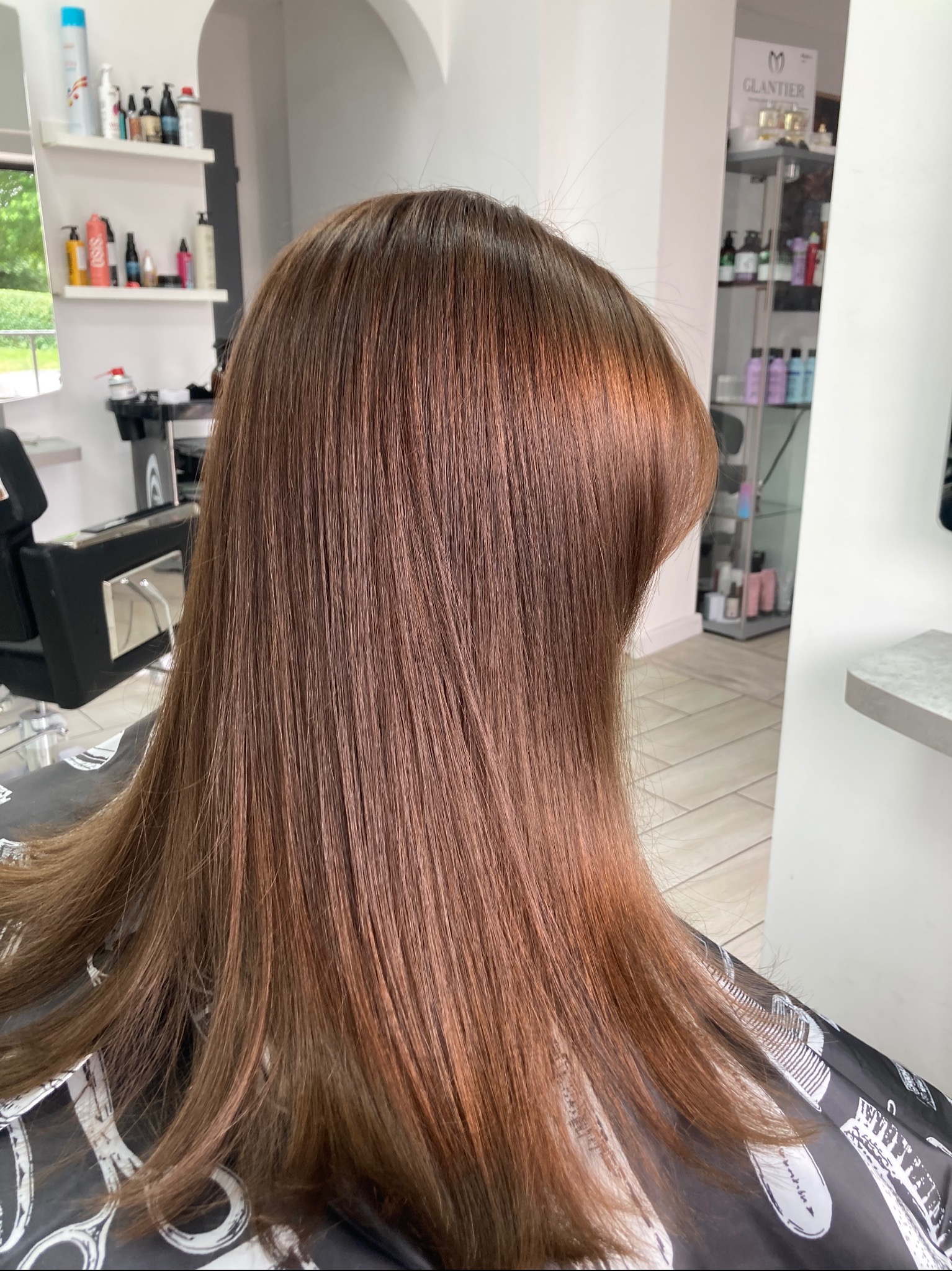 Świeżo ufarbowane, miedziano-brązowe, proste włosy klientki w salonie fryzjerskim, widoczne odbicie światła na grzywce, peleryna fryzjerska z białymi wzorami.