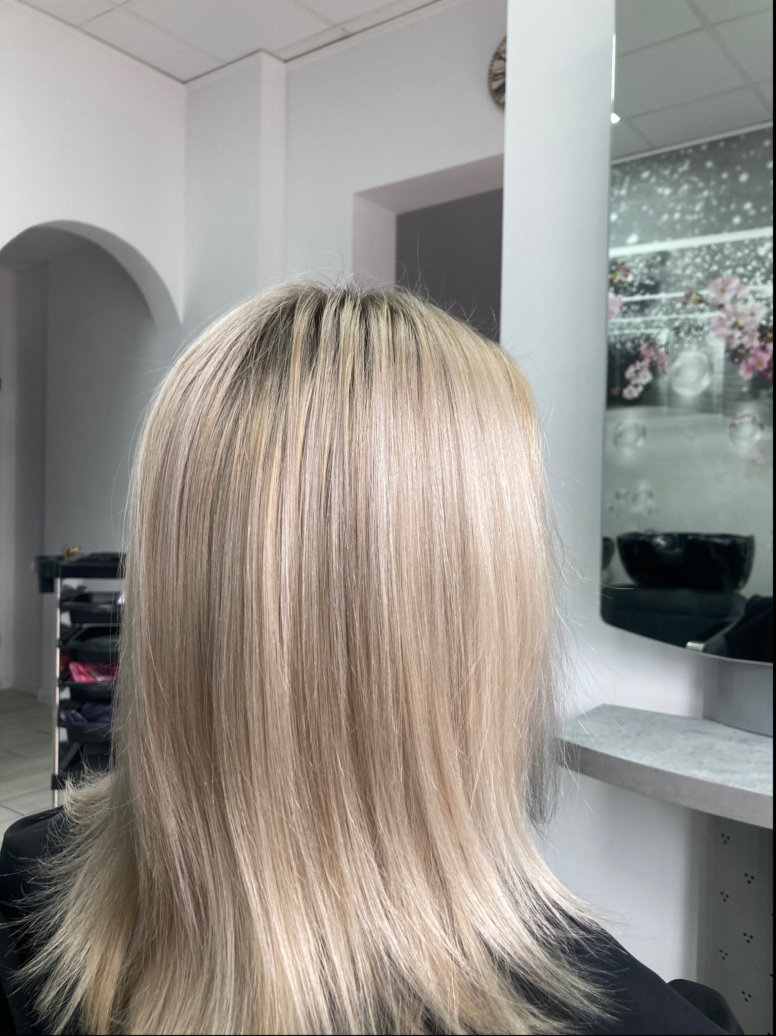 Tył głowy kobiety z włosami w odcieniach blondu, widoczne ciemniejsze odrosty, w tle jasne wnętrze salonu z elementami dekoracyjnymi.