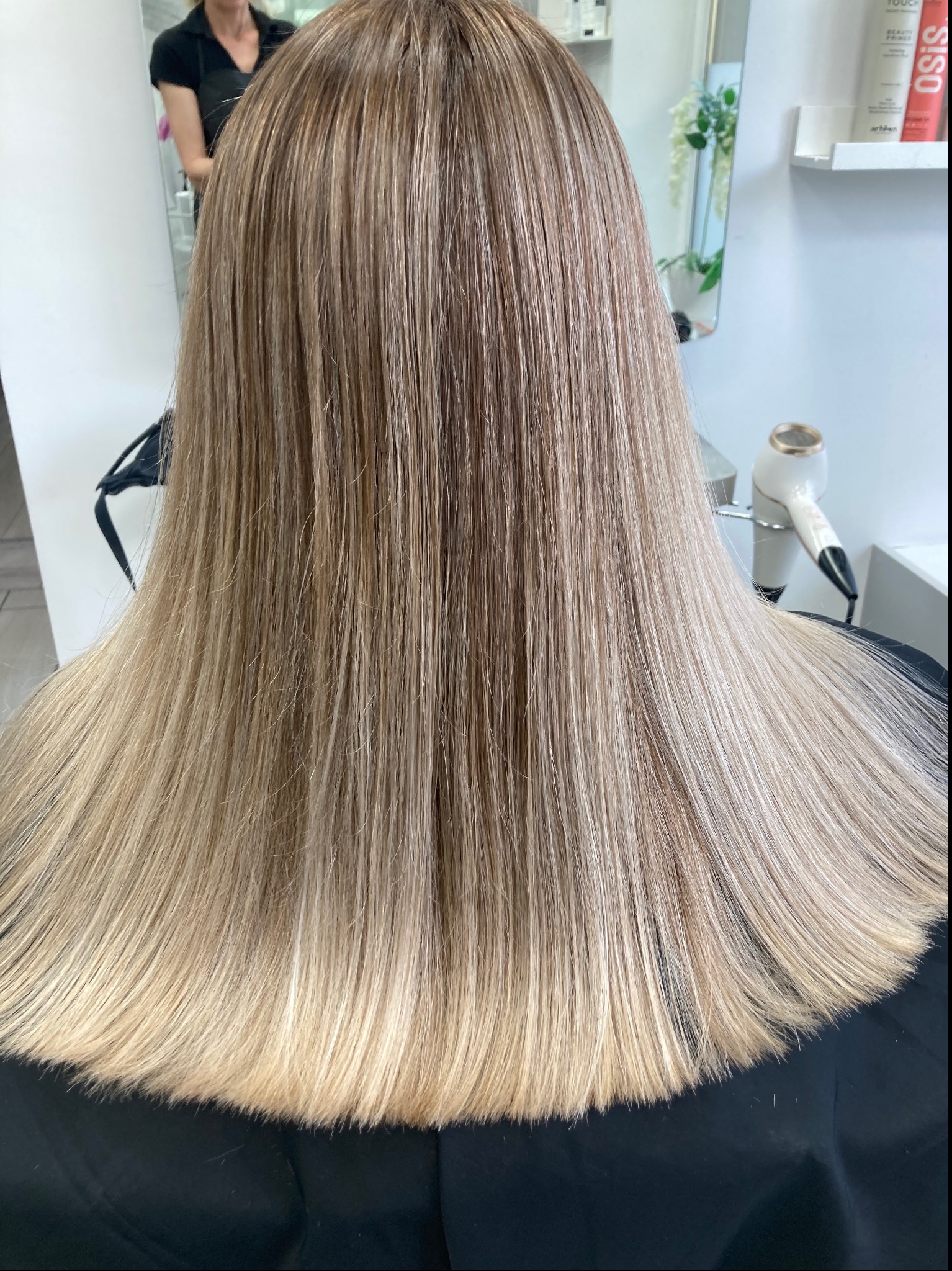 Długie, proste włosy z koloryzacją sombre, przechodzące od ciemniejszego blondu u nasady do jaśniejszego na końcach, widoczne z tyłu, w tle suszarka i lustro.