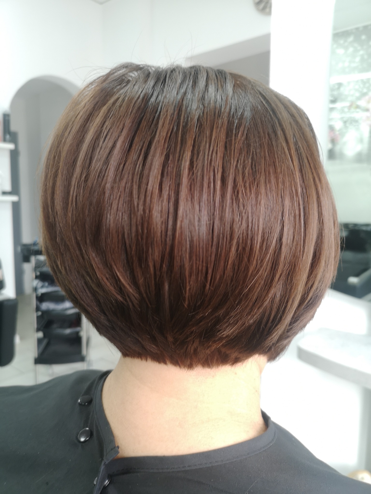 Krótkie, brązowe włosy ułożone w zaokrąglony bob, widoczne z tyłu głowy, z delikatnym cieniowaniem na końcach, na tle jasnego wnętrza salonu fryzjerskiego.