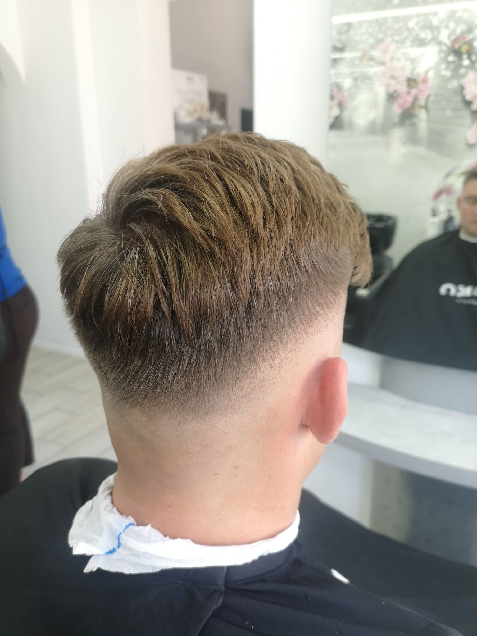 Męskie włosy po strzyżeniu z cieniowanym tyłem głowy, widoczny w lustrze fryzjer w czarnej pelerynie, białe ściany i dekoracje kwiatowe w tle.