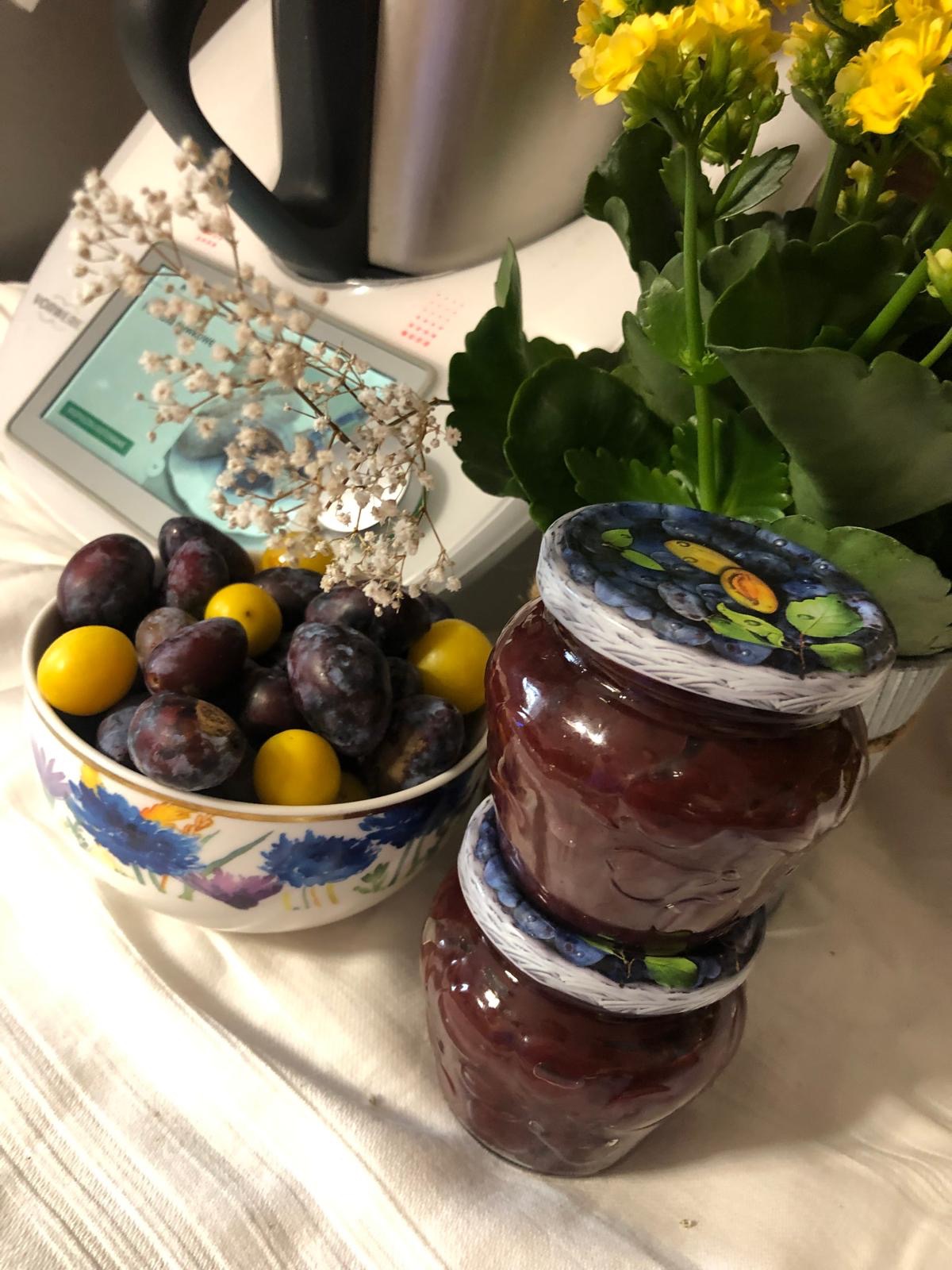 Domowe przetwory śliwkowe w słoikach z ręcznie malowanymi wieczkami, śliwki w miseczce i kwiaty na stole. W tle robot kuchenny.