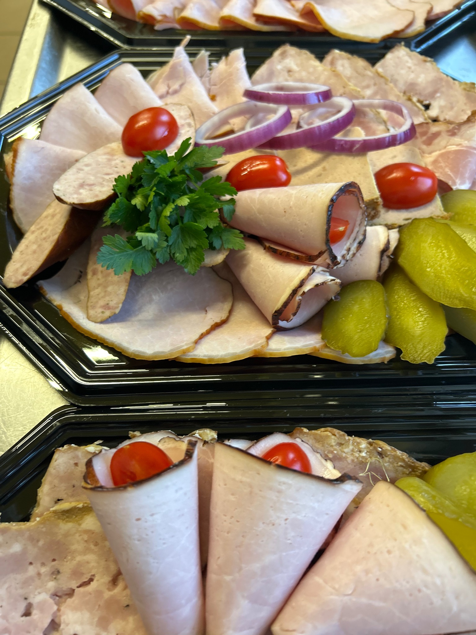 Elegancko ułożona taca z wędlinami, dekorowana pomidorkami cherry, ogórkami konserwowymi, czerwoną cebulą i zieloną pietruszką, gotowa do serwowania na przyjęciu.