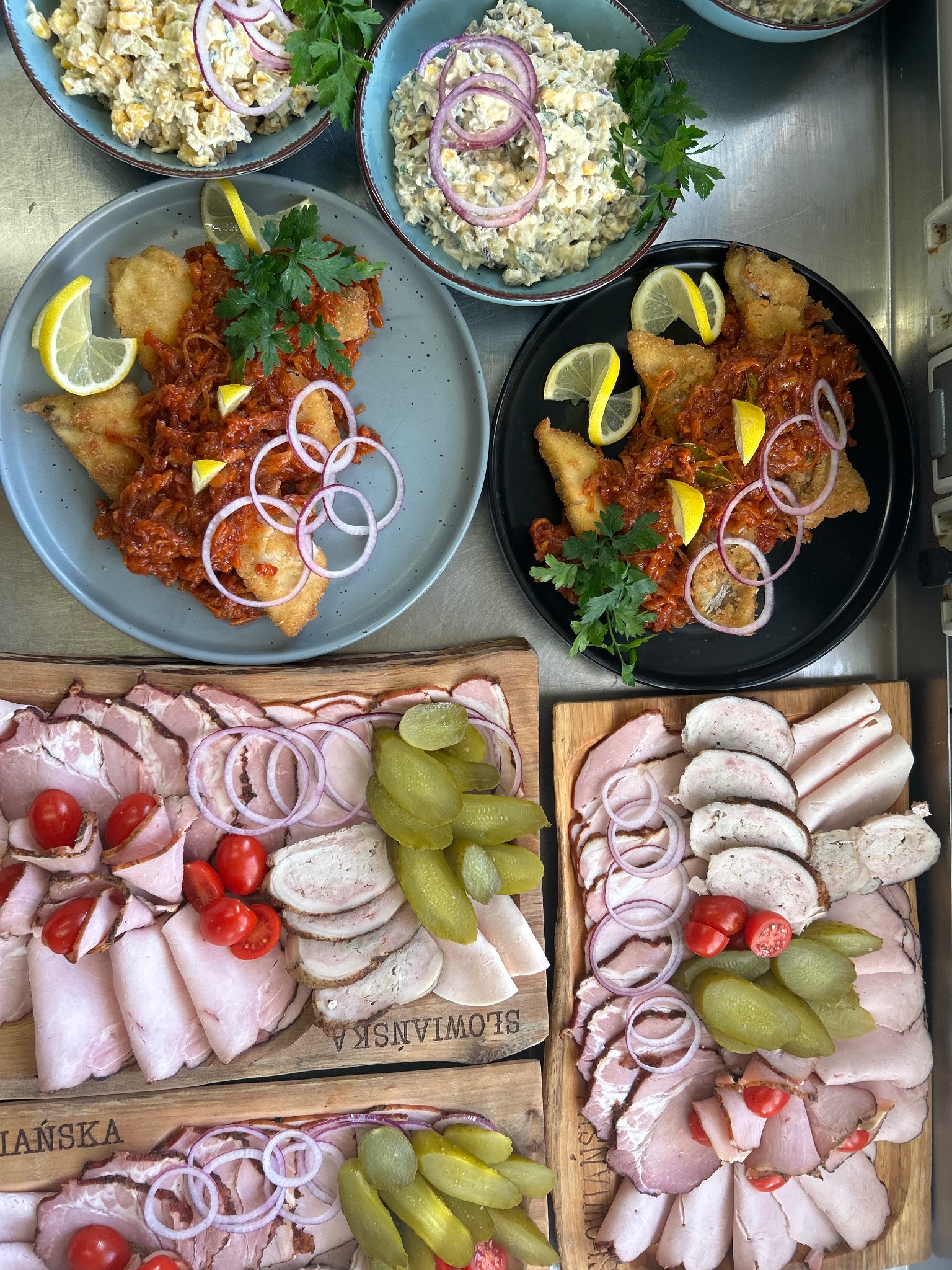 Bogato zastawiony stół cateringowy: ryba w sosie, sałatki w miskach i deski wędlin z dodatkami, tworzące apetyczną kompozycję.