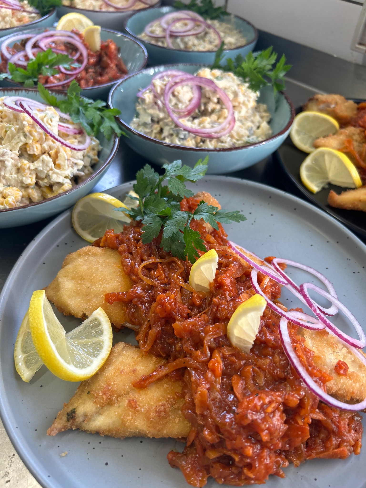 Elegancko podane danie: smażona ryba z sosem pomidorowym, cytryną i czerwoną cebulą na szarym talerzu. W tle miski z sałatkami, całość w jasnym oświetleniu.