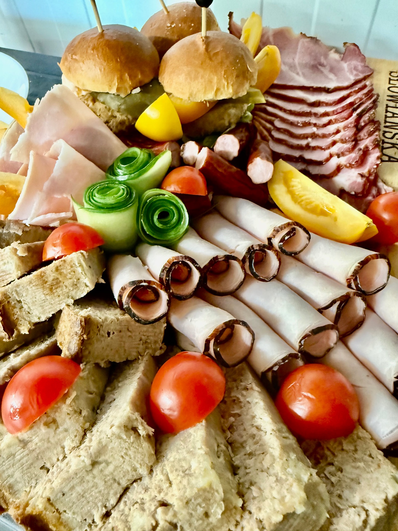 Bogato zaaranżowana deska przekąsek: mini burgery, wędliny w rolkach, plastry szynki, pomidorki koktajlowe, ogórki w rozetki i pieczywo. Idealna propozycja na catering.