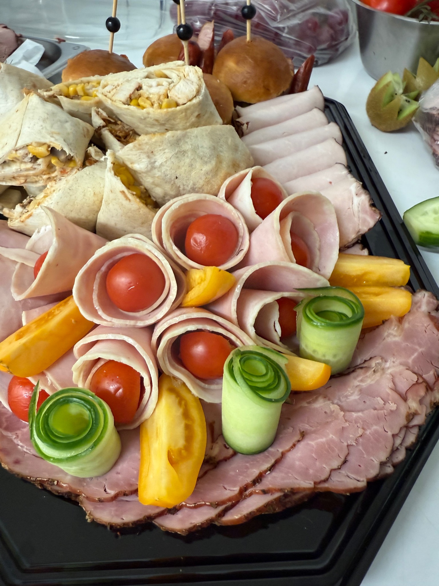 Elegancka taca cateringowa z roladkami szynki z pomidorkami, tortillami z kurczakiem i kukurydzą, mini burgerami i dekoracjami z ogórka i kiwi.