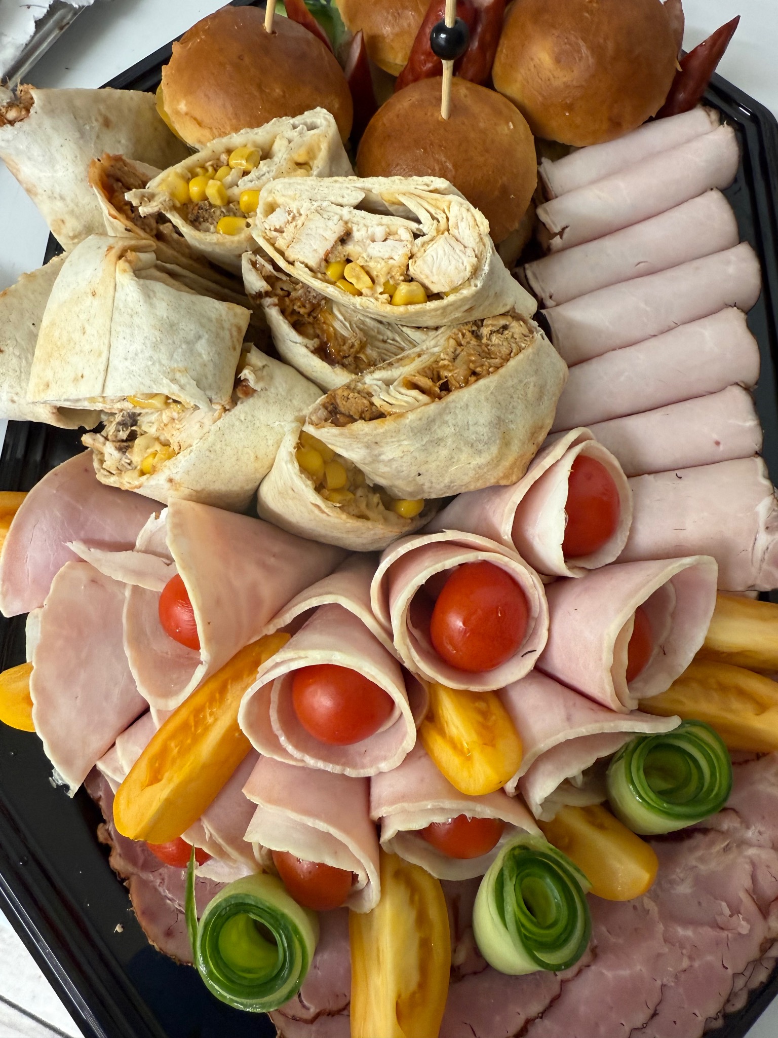 Dekoracyjna taca cateringowa z roladkami szynkowymi z pomidorkami, wrapami z kurczakiem i kukurydzą, mini burgerami i plastrami wędlin na czarnym talerzu.