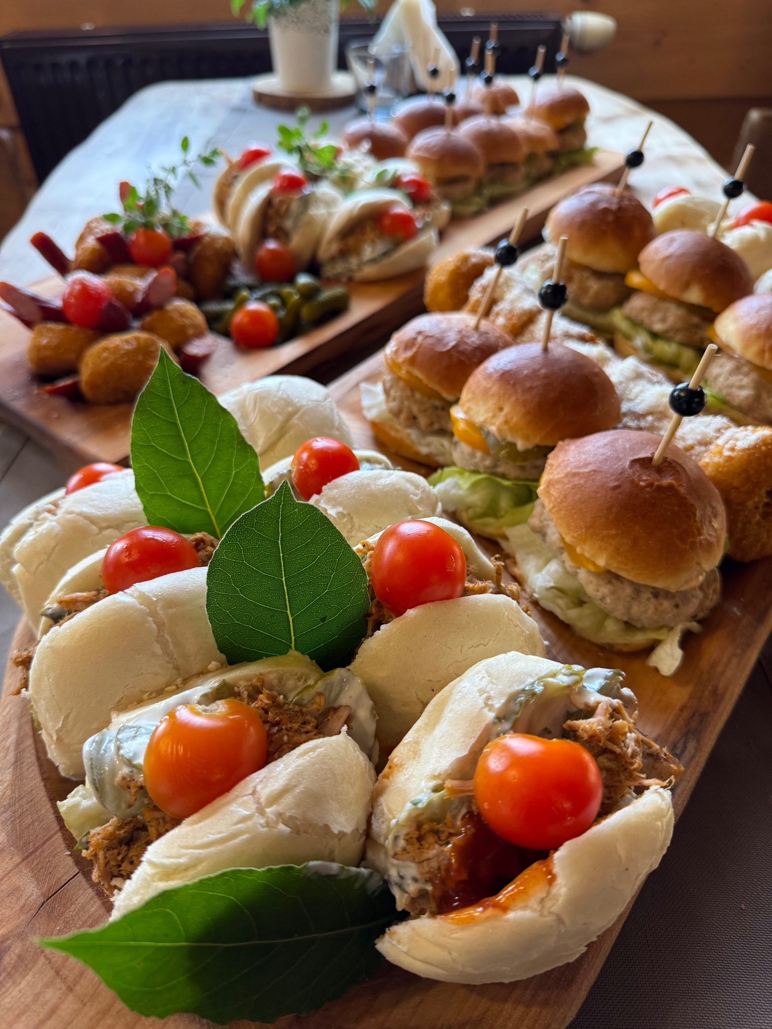 Drewniana deska z mini burgerami, kanapeczkami i przekąskami, udekorowana listkami i pomidorkami koktajlowymi. Elegancki catering na imprezę firmową.
