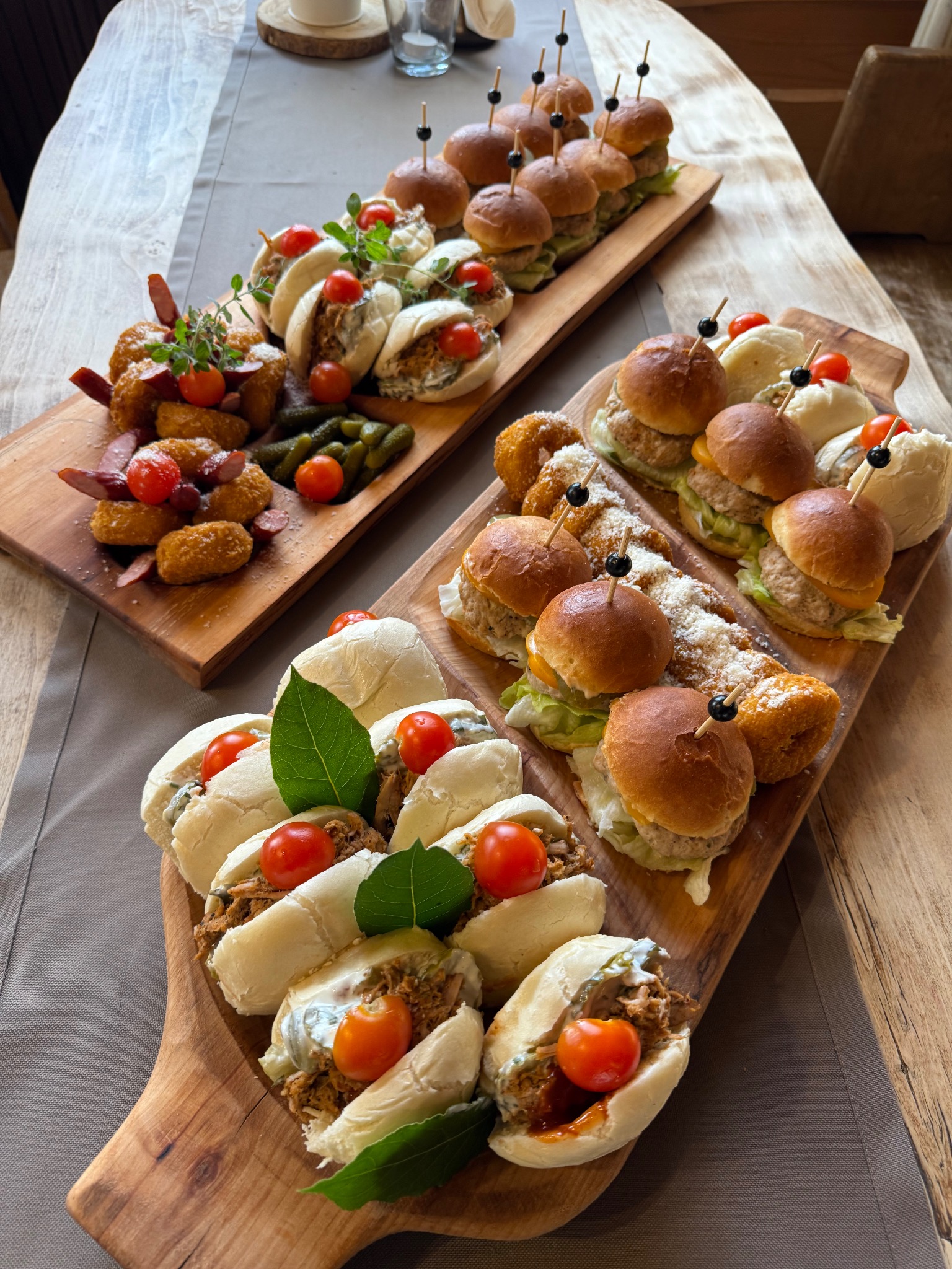 Dwie drewniane deski z mini burgerami, kanapeczkami z pomidorkami cherry, krokietami i ogórkami konserwowymi, ułożone na stole z szarą serwetą.