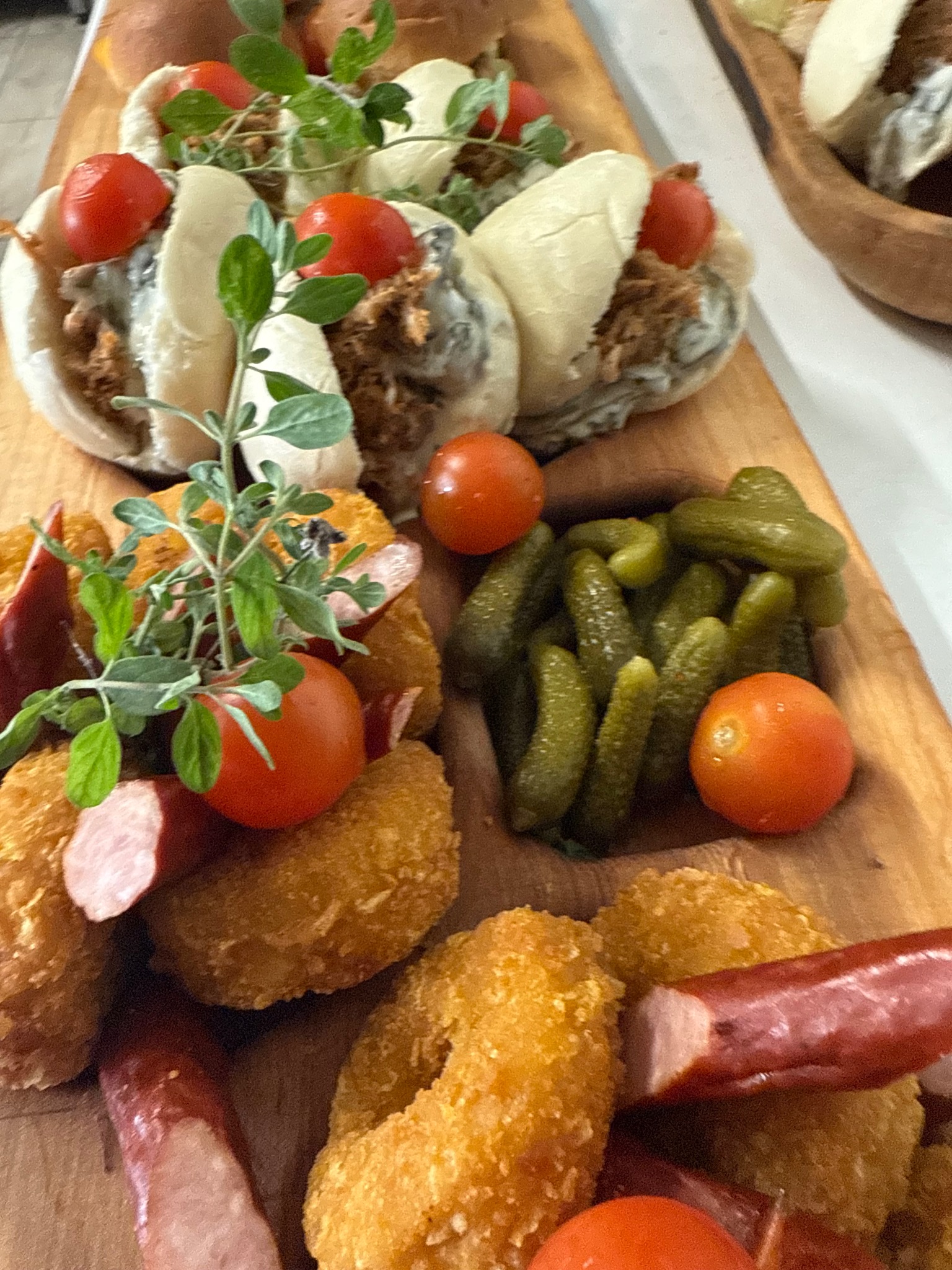 Deska z przekąskami: mini bułeczki z mięsem, ogórki konserwowe, krążki cebulowe, kiełbasa i pomidorki cherry, udekorowane świeżymi ziołami.