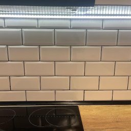 Płytka PL - Ściana w kuchni wyłożona białymi, podłużnymi płytkami ceramicznymi typu metro z ciemną fugą, oświetlona listwą LED pod szafką, widoczny fragment blatu i płyty indukcyjnej.