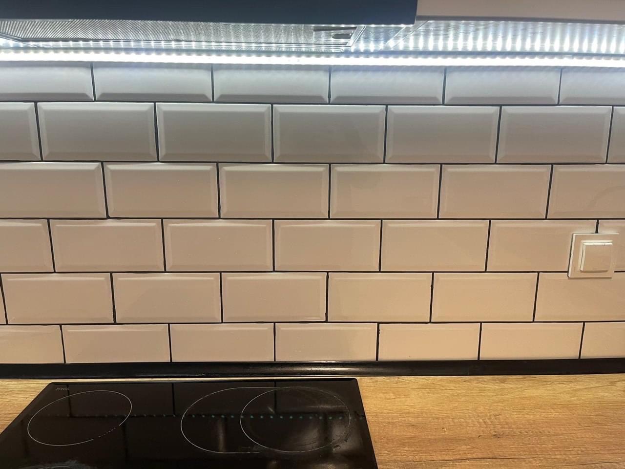 Ściana w kuchni wyłożona białymi, podłużnymi płytkami ceramicznymi typu metro z ciemną fugą, oświetlona listwą LED pod szafką, widoczny fragment blatu i płyty indukcyjnej.