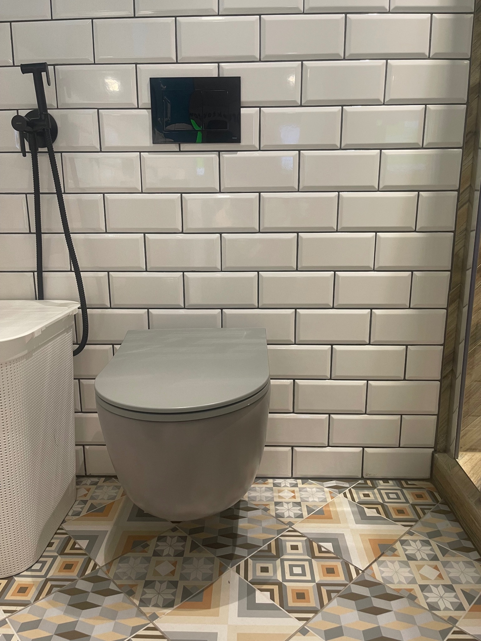 Nowoczesna łazienka z białą, cegiełkową glazurą, podwieszaną miską WC w kolorze szarym, czarną armaturą bidetową i wzorzystą, kolorową podłogą z płytek.