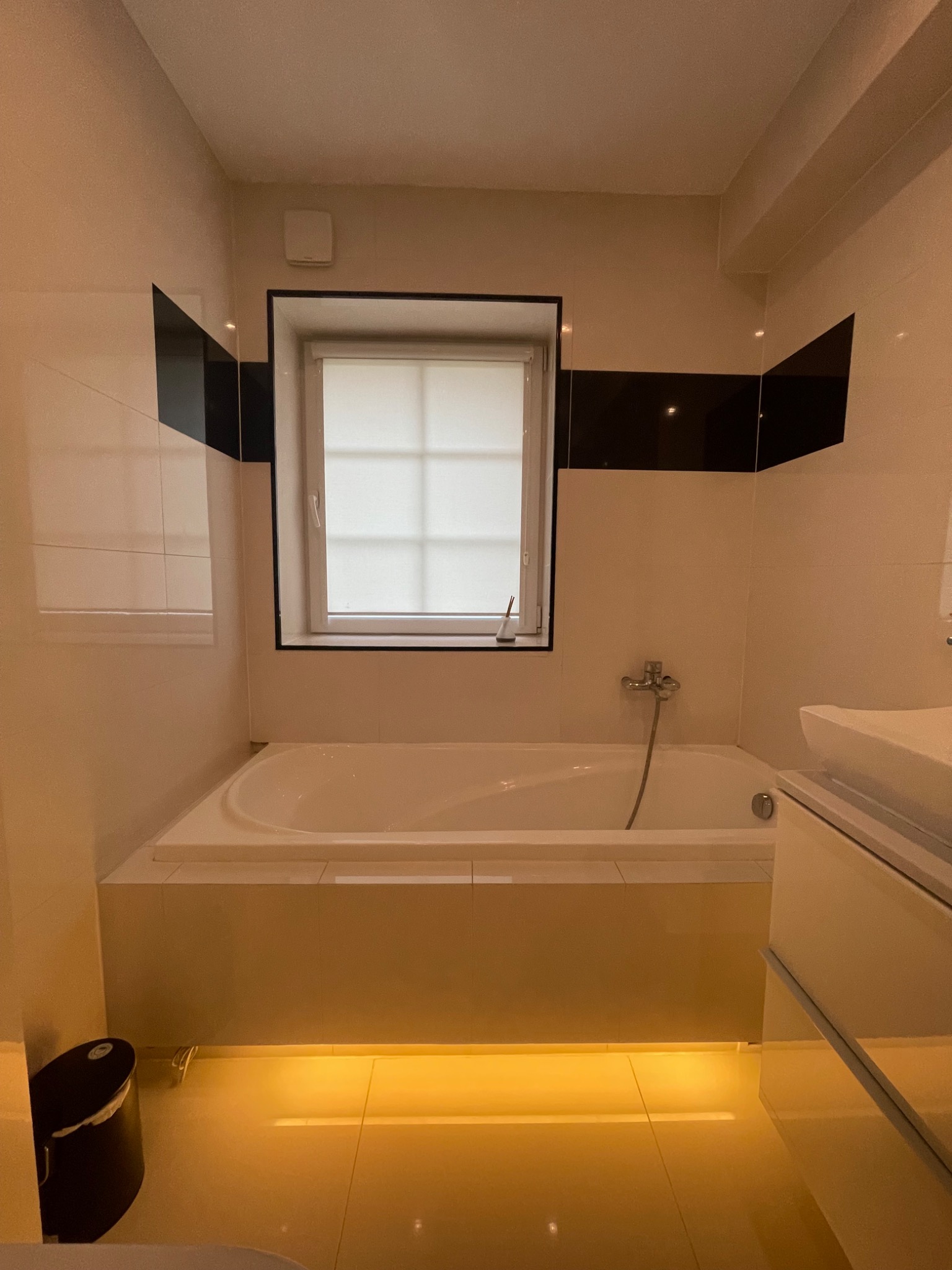 Nowoczesna łazienka z białą wanną obudowaną płytkami, czarnymi pasami płytek na ścianie, oświetleniem LED pod wanną i minimalistyczną umywalką.