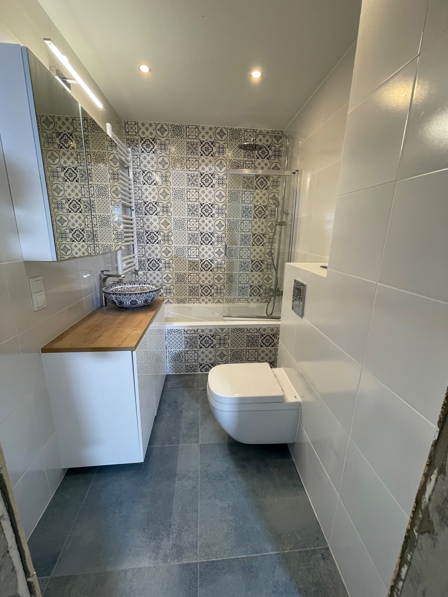 Nowoczesna łazienka po remoncie: białe płytki ścienne, szara podłoga, wisząca toaleta, umywalka w stylu retro na drewnianym blacie i dekoracyjne płytki nad wanną ze szklanym parawanem.