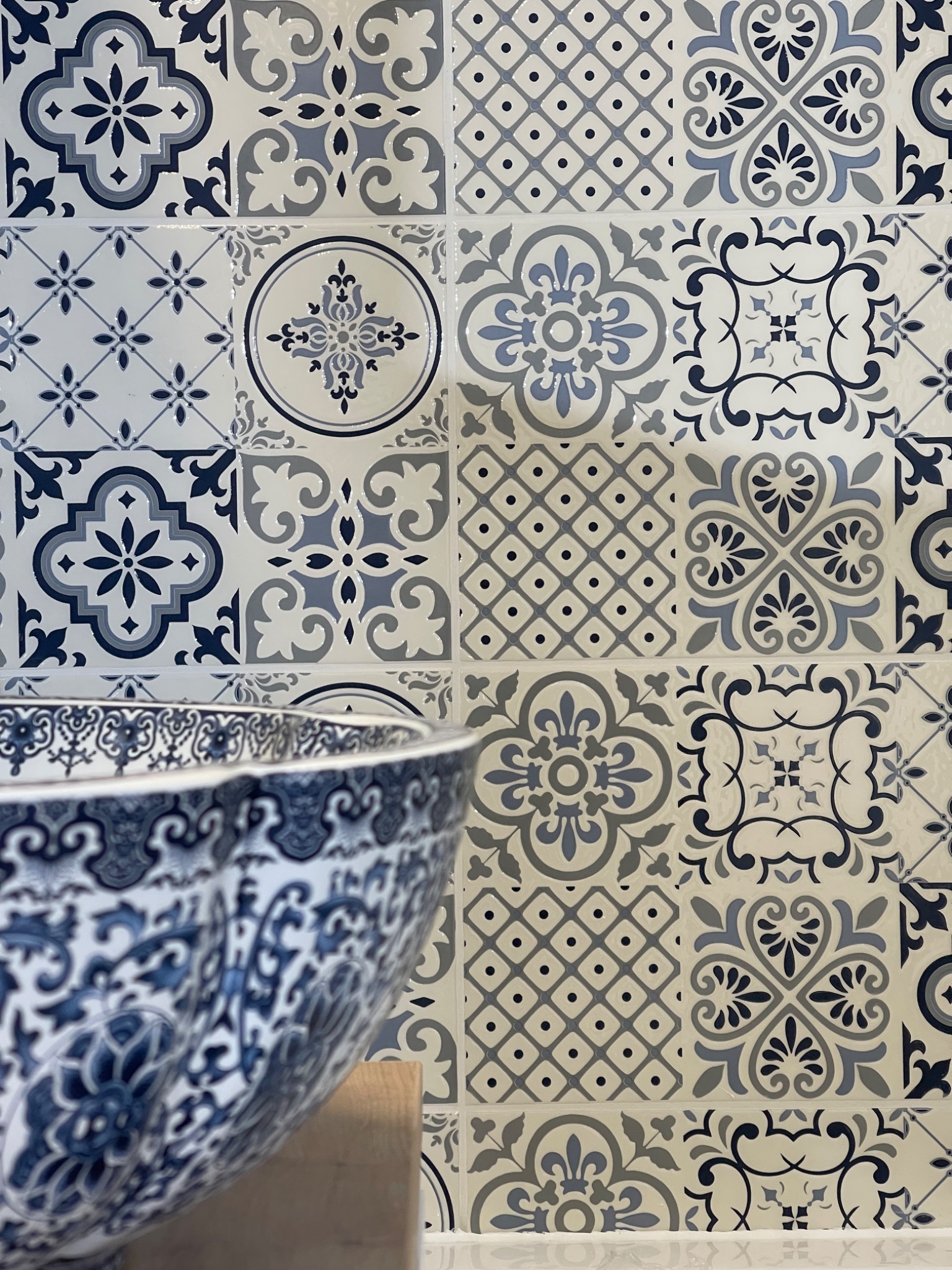 Błękitno-szare płytki dekoracyjne z motywami kwiatowymi i geometrycznymi na ścianie łazienki, obok umywalka z podobnym wzorem.