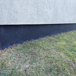 Usługi budowlane&stolarskie-Domino - Ocieplenie fundamentu budynku z widoczną warstwą izolacyjną w kolorze grafitowym, stykającą się z trawnikiem.