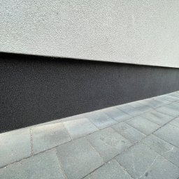 Usługi budowlane&stolarskie-Domino - Dolna część ściany budynku pokryta czarnym, chropowatym tynkiem mozaikowym, kontrastująca z szarą, gładką górną częścią ściany i szarą kostką brukową chodnika.