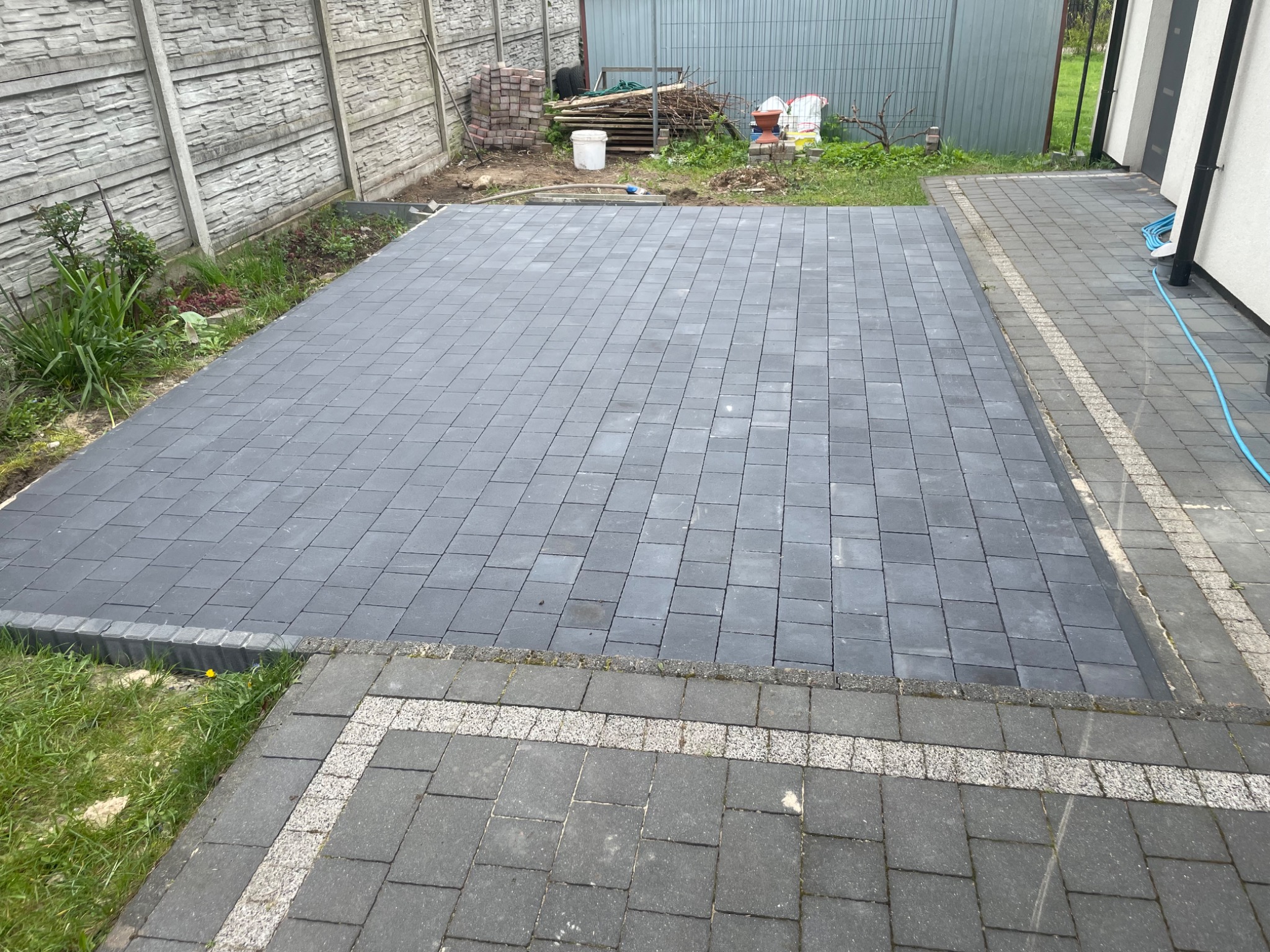 Nowo ułożona, szara kostka brukowa na posesji, obrzeża z ciemniejszej kostki, widoczny fragment ogrodu z chwastami i betonowe ogrodzenie w tle.