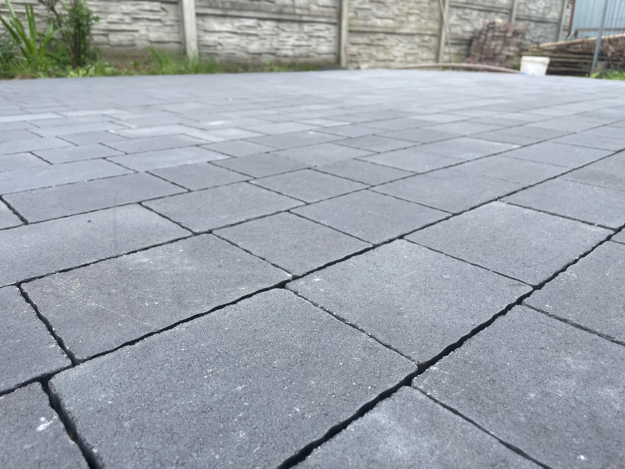 Szara kostka brukowa ułożona na posesji, widok z niskiej perspektywy, na tle betonowego ogrodzenia i składowanych elementów budowlanych.