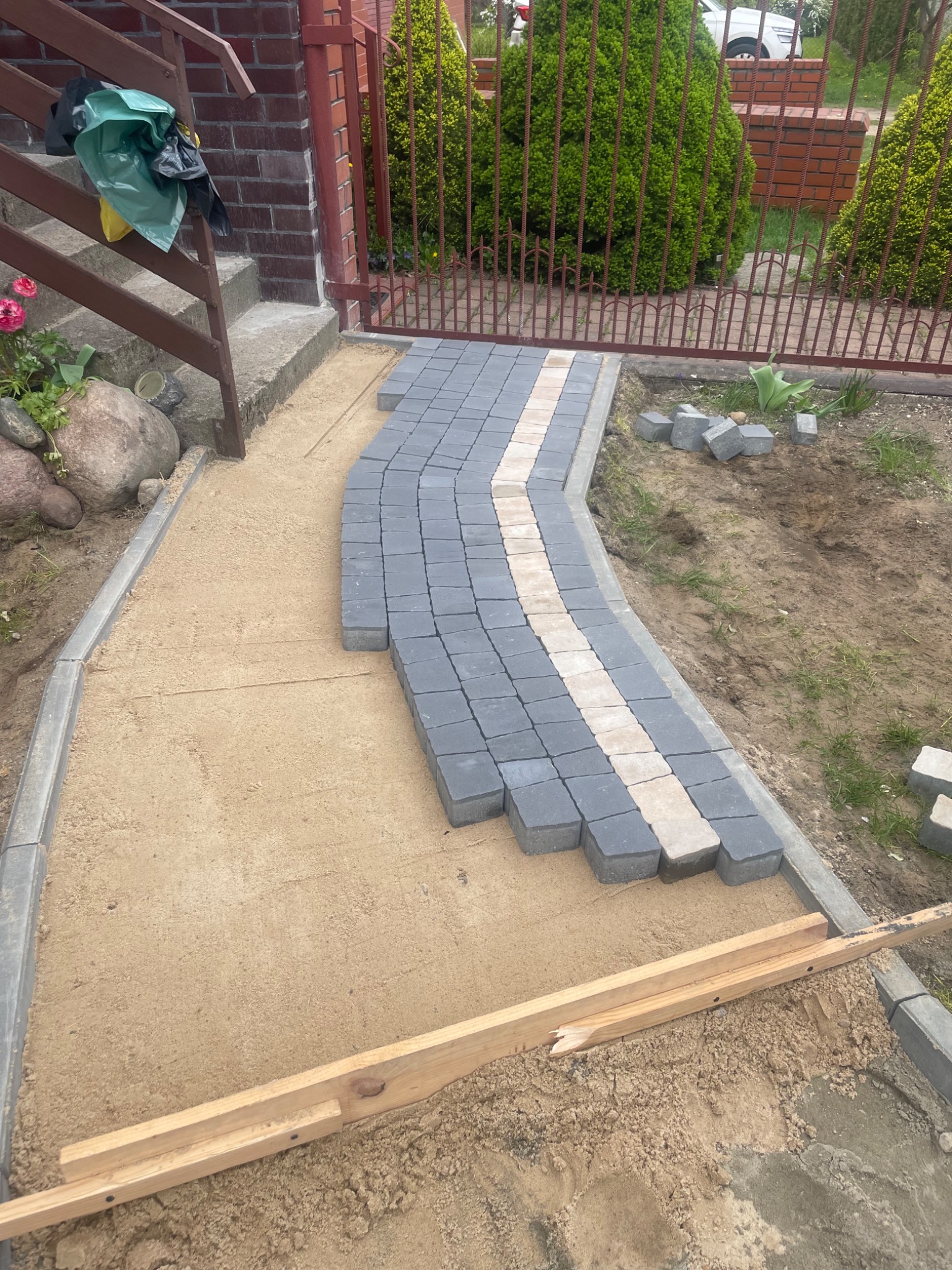 Wykonywanie ścieżki z kostki brukowej w ogrodzie, częściowo ułożona z szarych i beżowych elementów, widoczny piasek, obrzeża i drewniana listwa, w tle brama i zieleń.