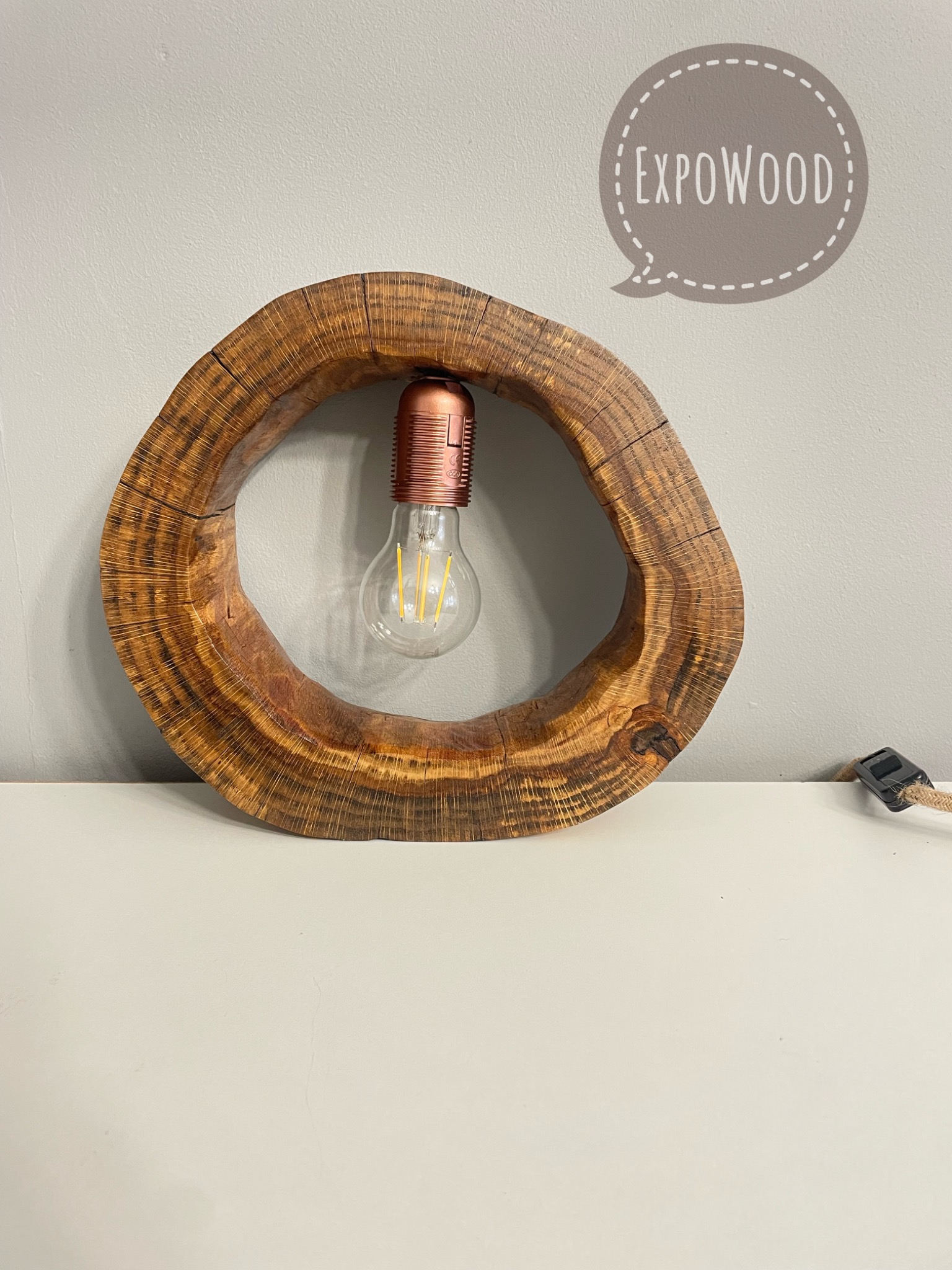 Lampa z plastra drewna z żarówką Edison, etykieta 'EXPOWOOD' na ścianie, kabel w oplocie jutowym.