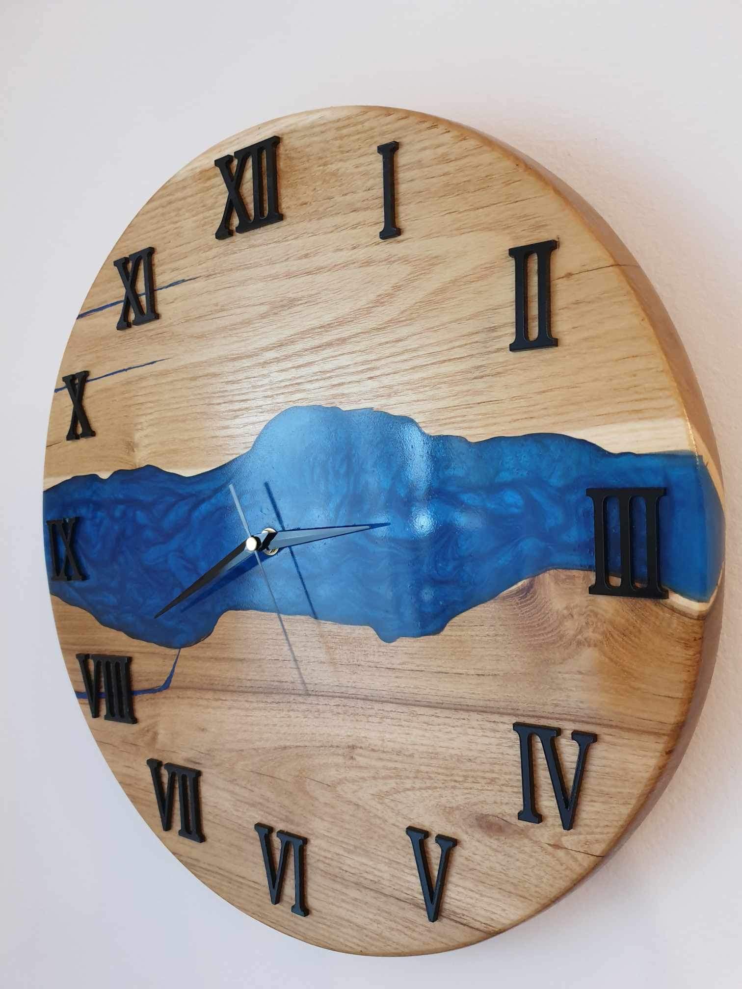 Okrągły zegar ścienny z jasnego drewna z czarnymi cyframi rzymskimi i wstawką z niebieskiej żywicy imitującą rzekę, wskazówki ustawione na godzinę 2:58.