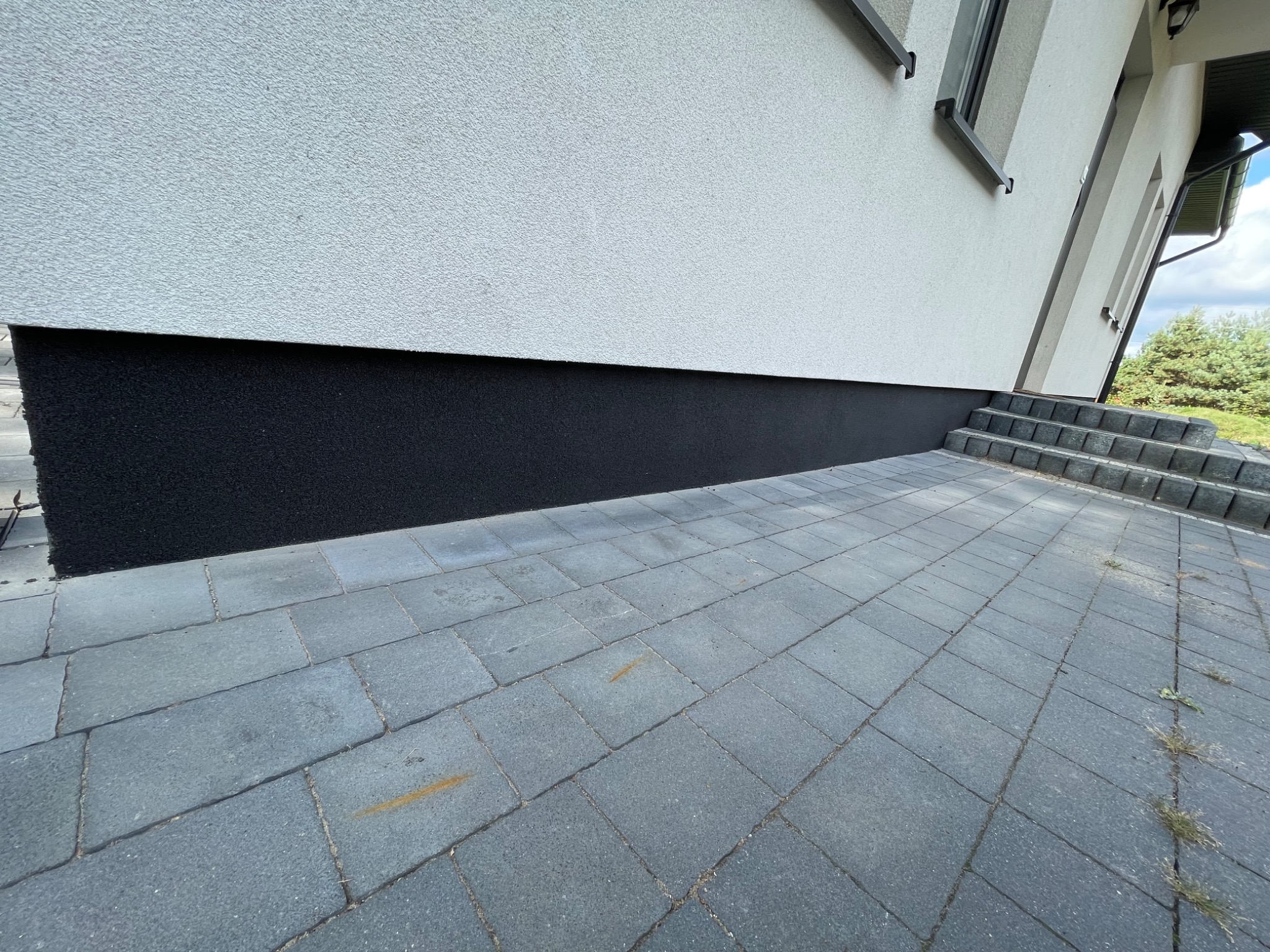 Elewacja budynku z kontrastowym, czarnym cokołem, wykończona jasnym tynkiem strukturalnym, widoczne schody wejściowe z kostki brukowej i fragment podjazdu.