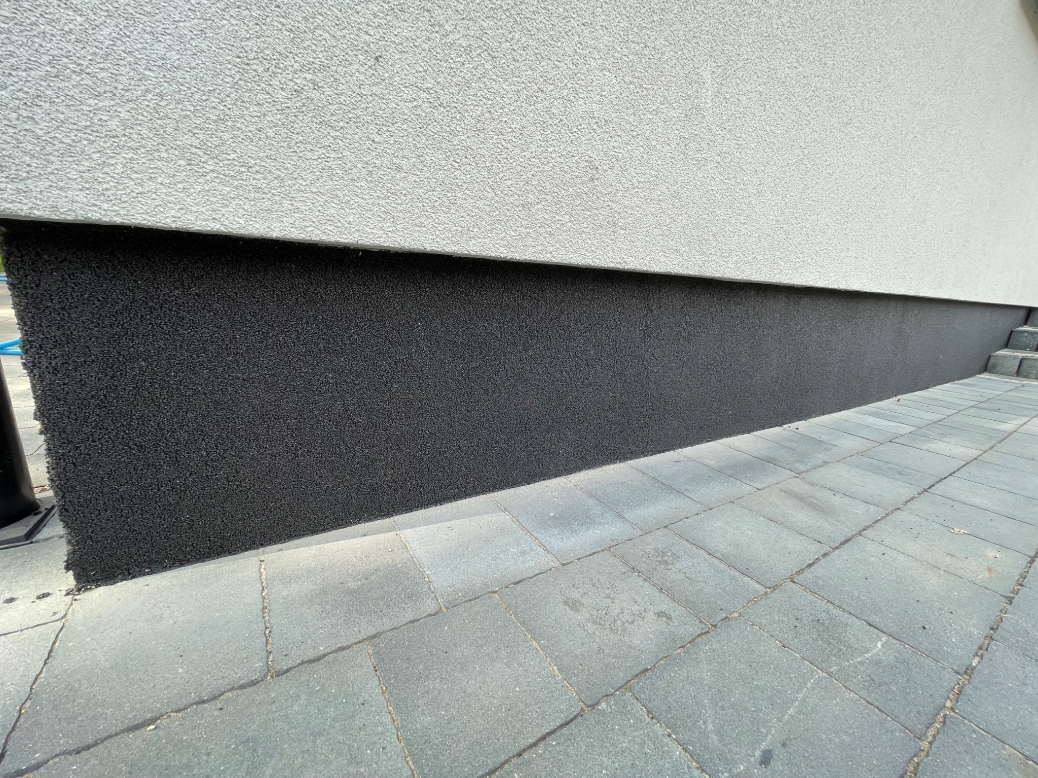 Dolna część ściany budynku pokryta czarnym, chropowatym tynkiem mozaikowym, kontrastująca z szarą, gładką górną częścią ściany i szarą kostką brukową chodnika.