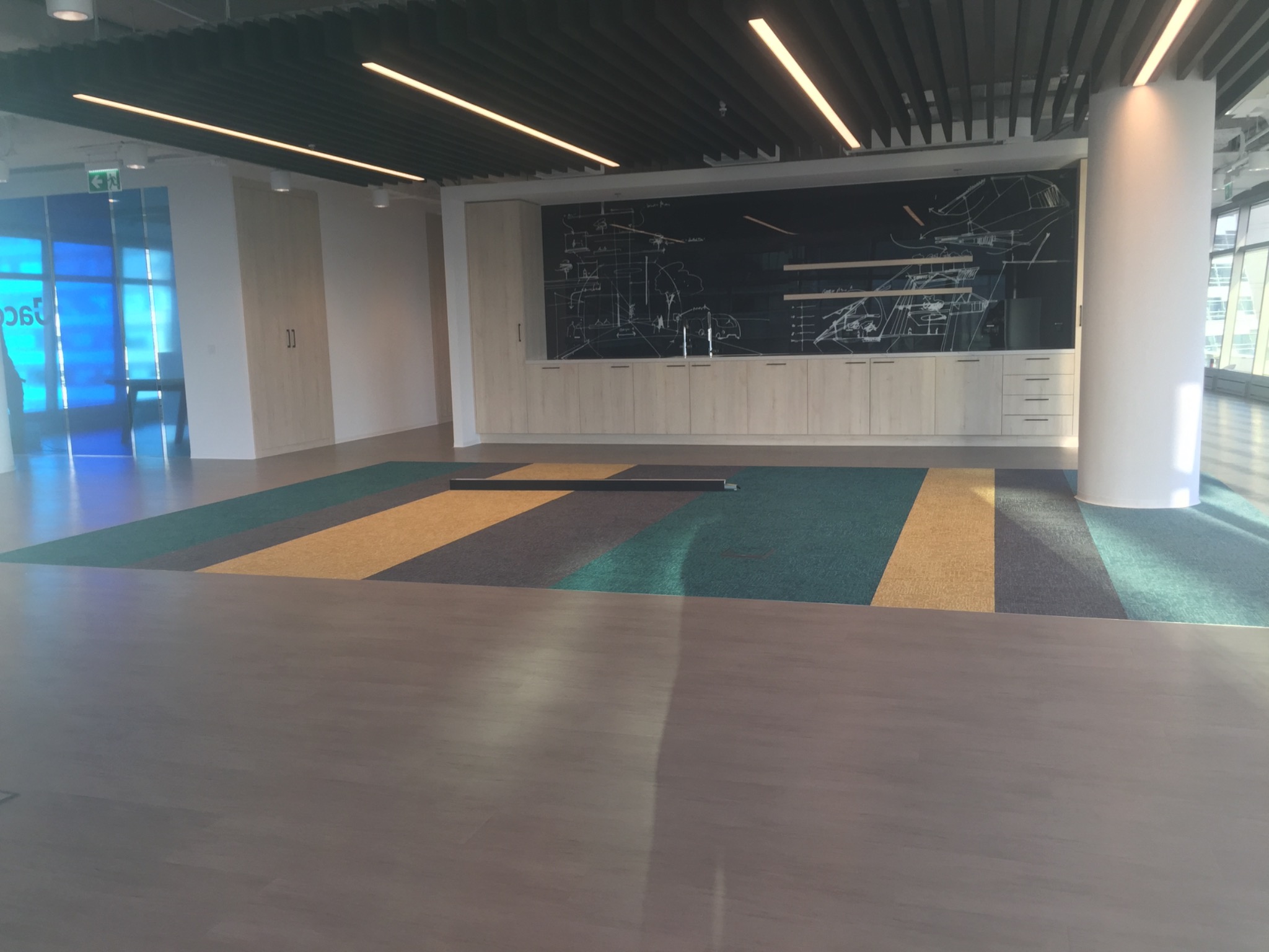 Przestronne wnętrze biurowe z drewnianą podłogą, wykładziną dywanową w pasy (zielony, żółty, szary) i zabudową meblową z tablicą z rysunkami technicznymi.