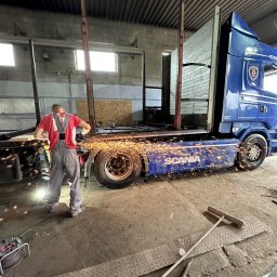Smal-Trans Jakub Smaluch - Spawanie ramy naczepy ciężarówki Scania w warsztacie, iskry rozbryzgują się spod tarczy szlifierki, pracownik w czerwonej koszulce i szarych spodniach roboczych, brudna podłoga warsztatu, widoczny...
