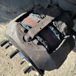 Brudny zacisk hamulcowy z widocznymi klockami i śrubami mocującymi, oświetlony z góry, z numerami seryjnymi na obudowie.