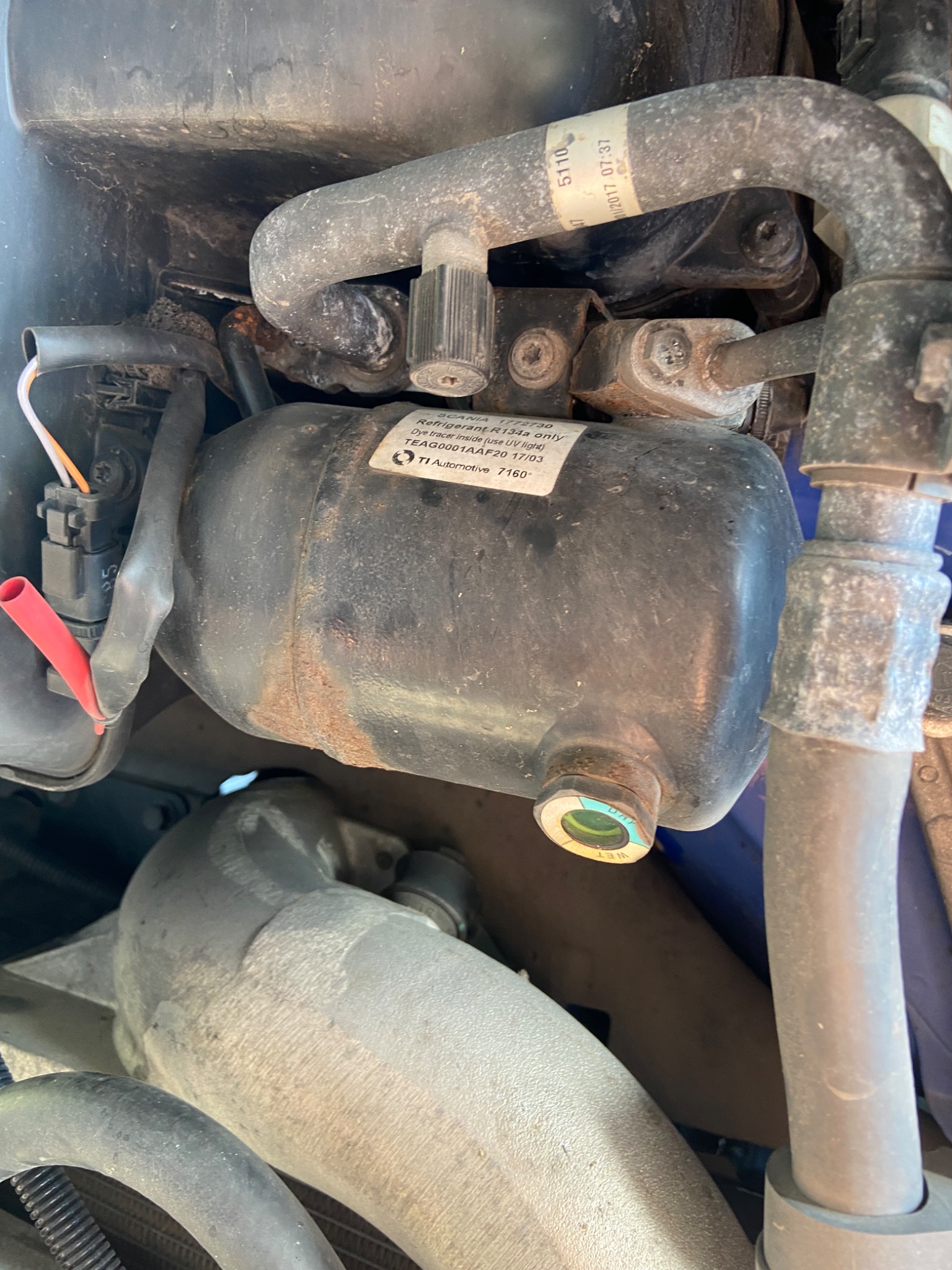 Zbliżenie na element układu klimatyzacji samochodowej z widocznymi przewodami, złączkami i zbiornikiem czynnika chłodniczego, z naklejką informacyjną i śladami korozji.