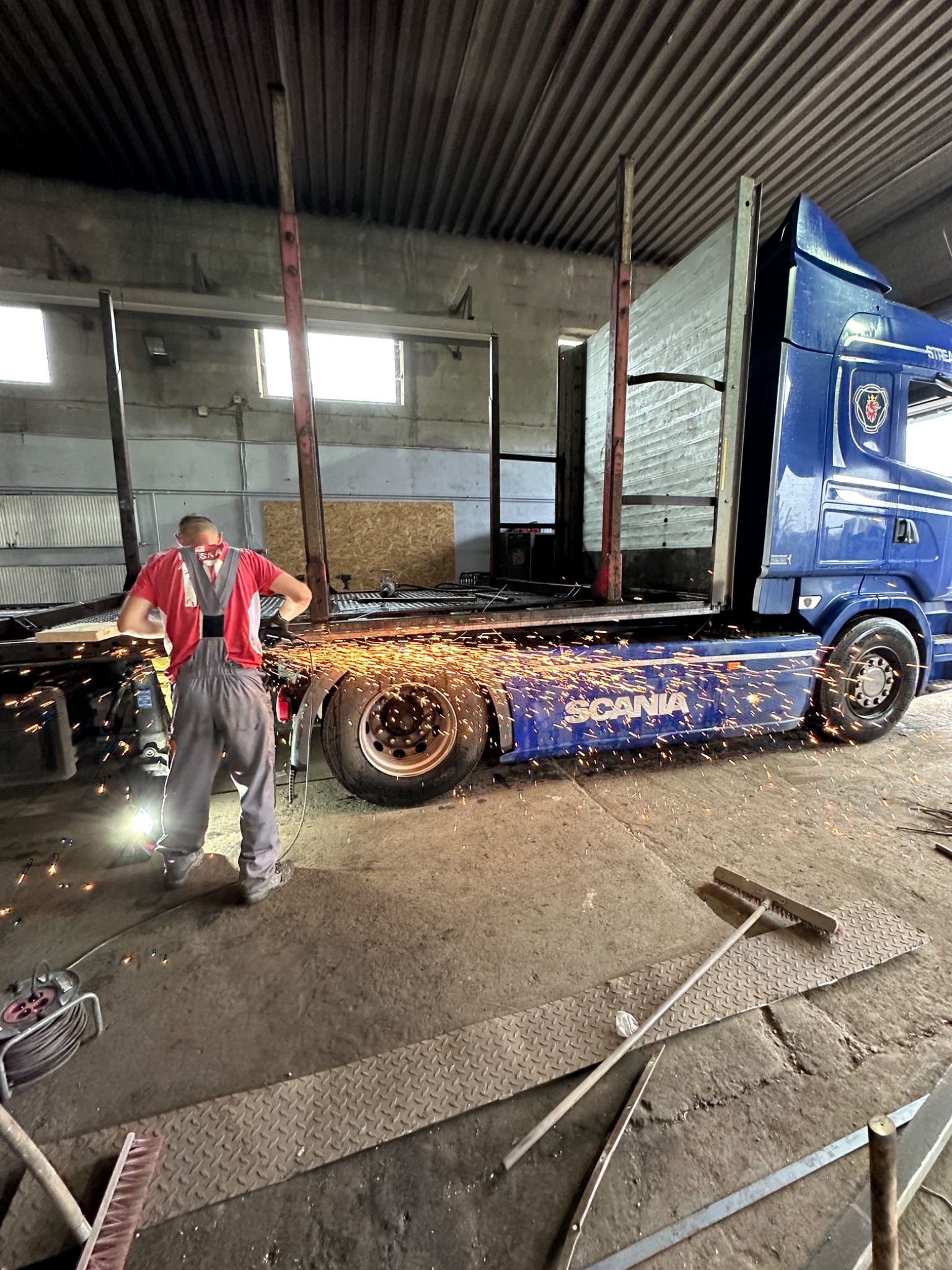 Spawanie ramy naczepy ciężarówki Scania w warsztacie, iskry rozbryzgują się spod tarczy szlifierki, pracownik w czerwonej koszulce i szarych spodniach roboczych, brudna podłoga warsztatu, widoczny...