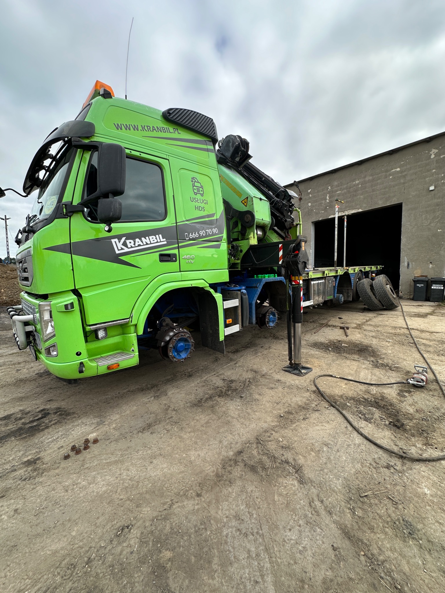 Zielony samochód ciężarowy z dźwigiem marki KRANBIL, z podniesioną osią i zdjętymi kołami po jednej stronie, stoi na placu przed budynkiem warsztatowym, widoczne narzędzia i śruby.