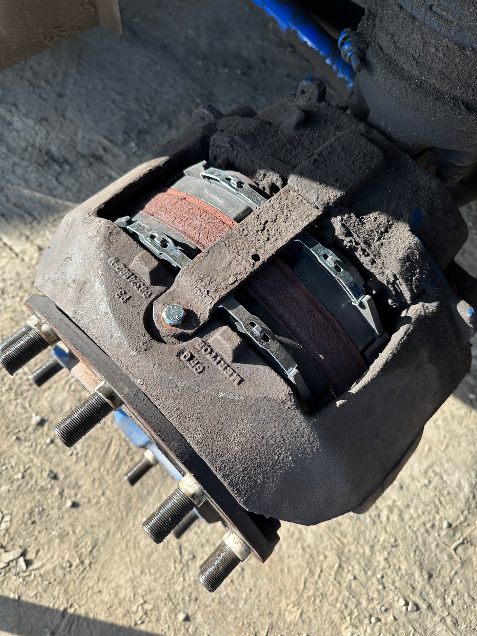 Brudny zacisk hamulcowy z widocznymi klockami i śrubami mocującymi, oświetlony z góry, z numerami seryjnymi na obudowie.